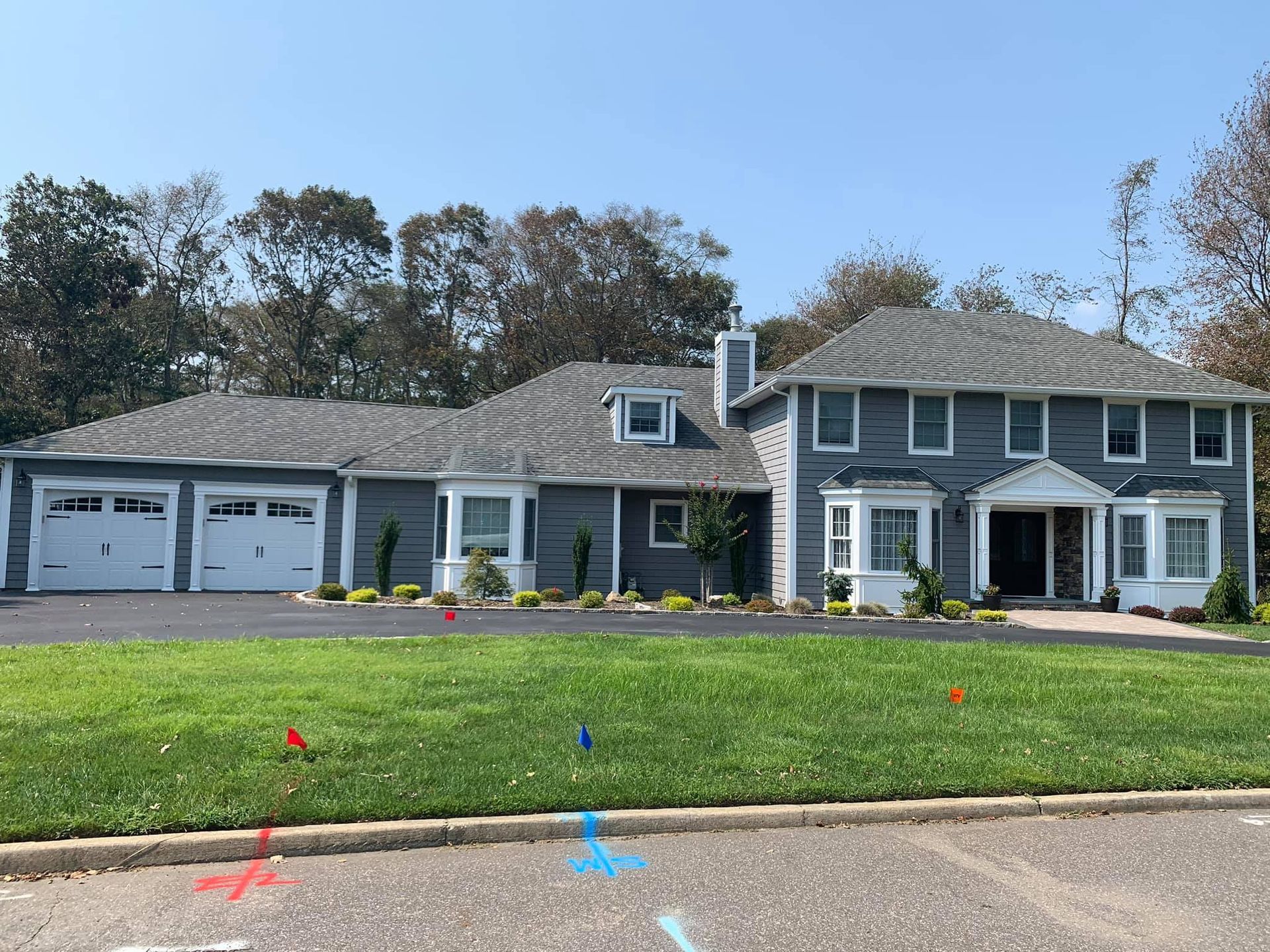 A two-story grey suburban house with a multi-car garage, a green front lawn, and colorful utility flags in the grass.