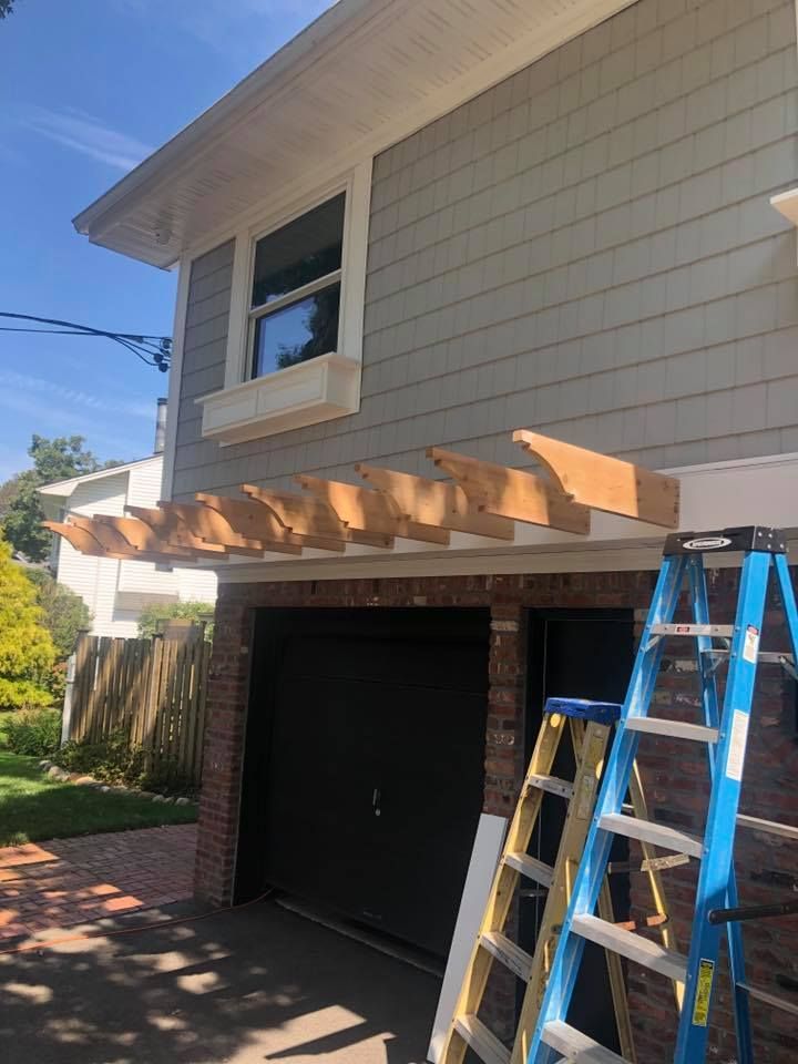 Unfinished wooden pergola beams mounted above a garage door on a house with grey siding and two ladders nearby.