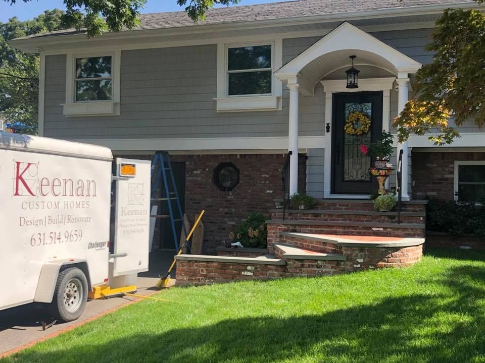 A Keenan Custom Homes trailer parked on a driveway in front of a gray-sided home with brick masonry and a porch entryway.