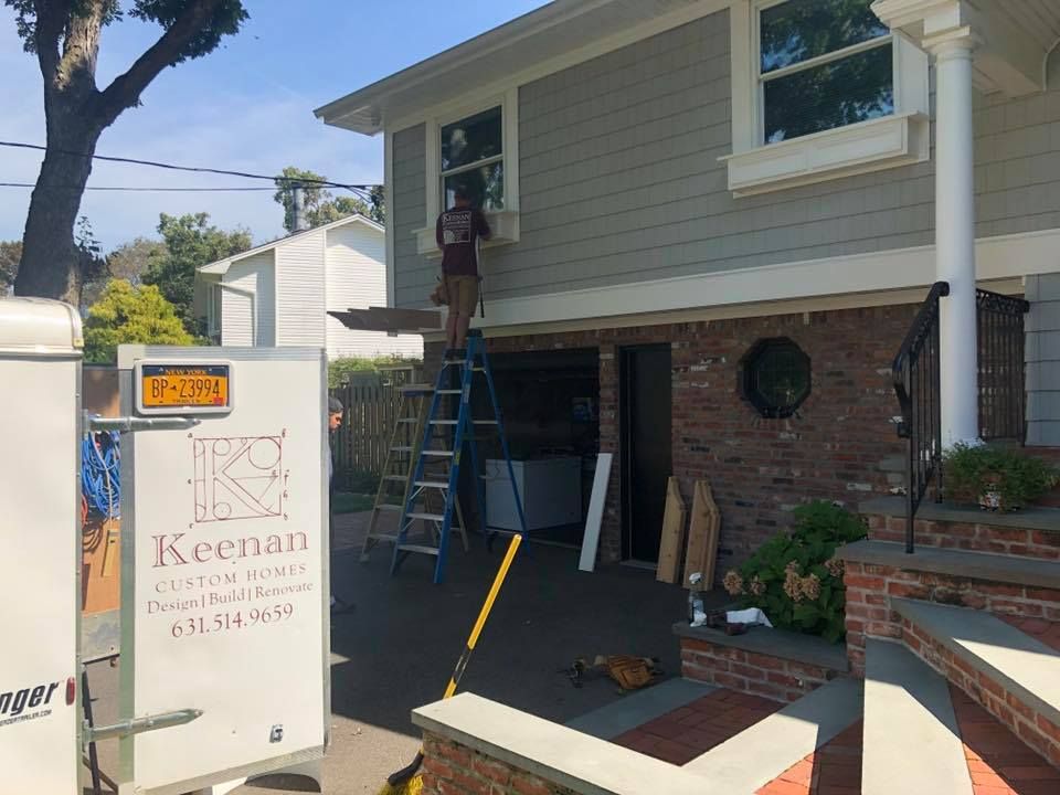A worker on a ladder installs a window on the upper story of a house with gray siding and a brick lower level.