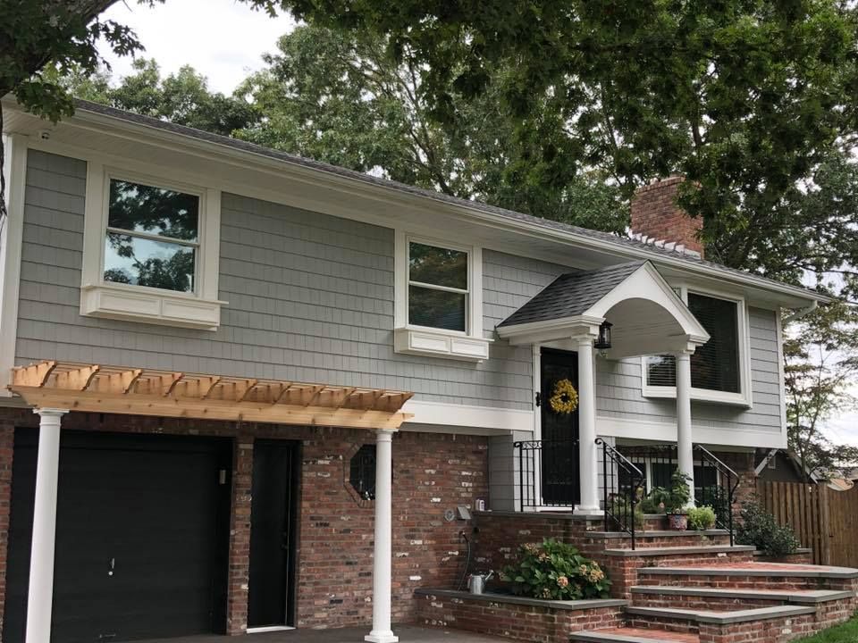 A two-story house with gray shingle siding, a red brick base, a black garage door, and a front porch with white columns.