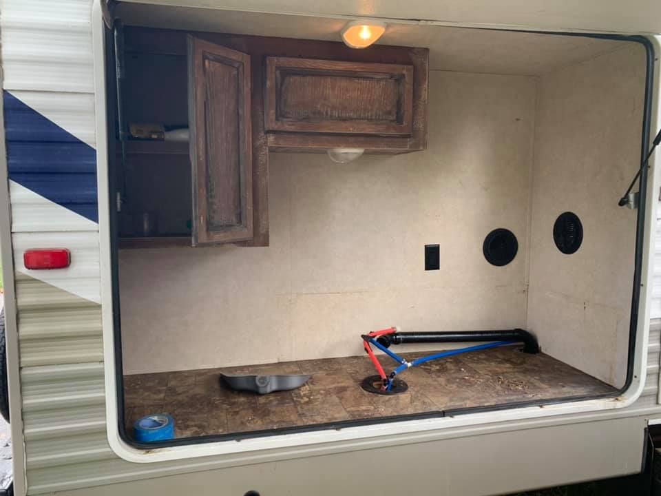 An open RV exterior storage compartment with a cabinet, light, power outlet, and exposed water lines on the floor.