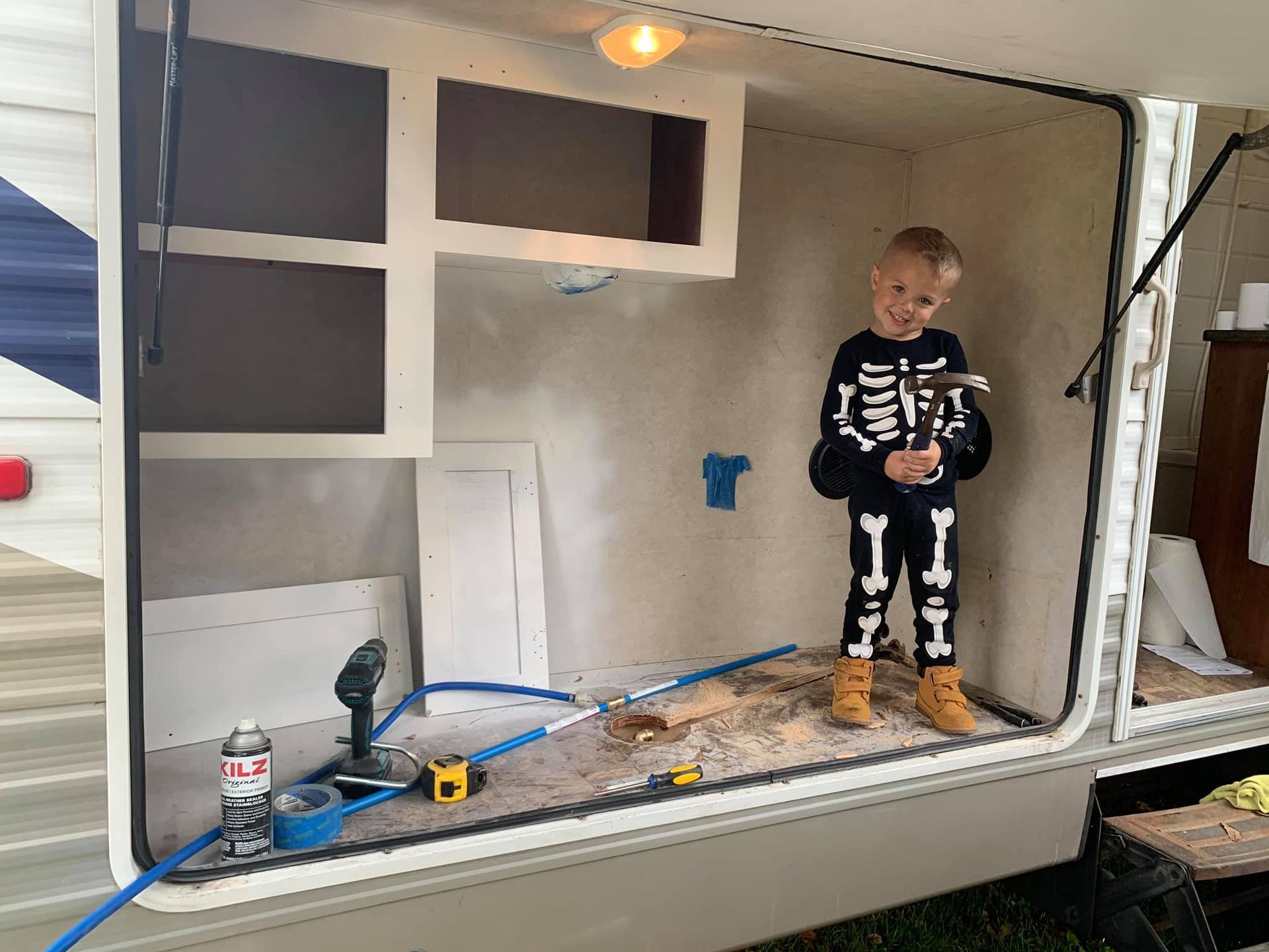 A child in a skeleton costume holding a hammer stands inside an unfinished, light-colored storage bay of a camper.
