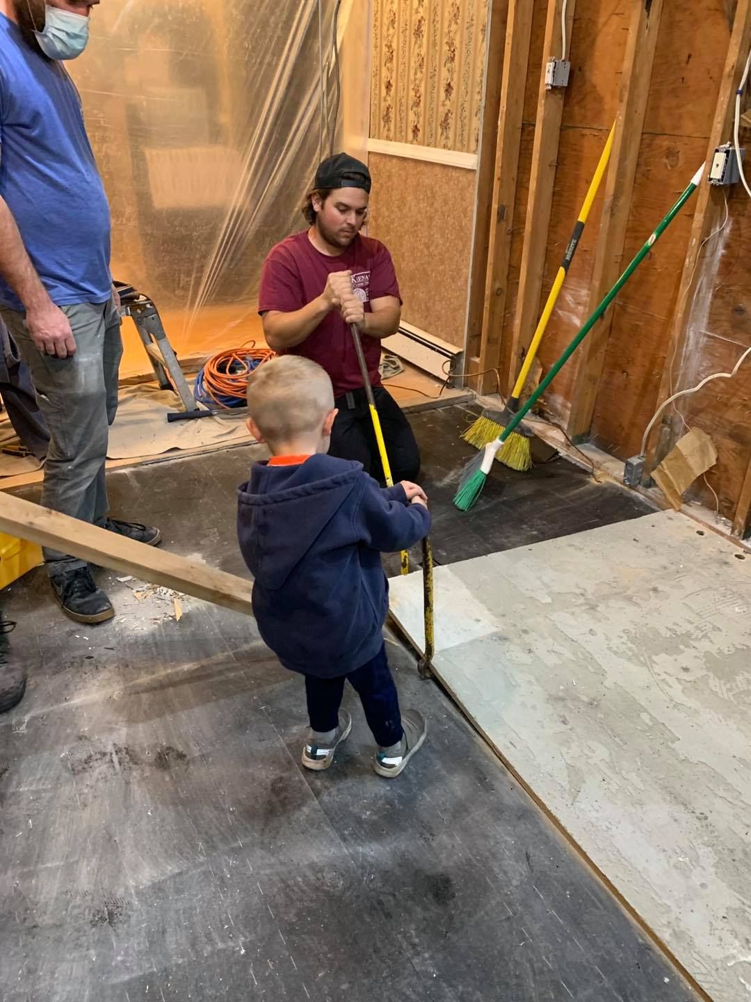 A person in a maroon shirt and a young child use a tool to work on the floor in a room undergoing construction.