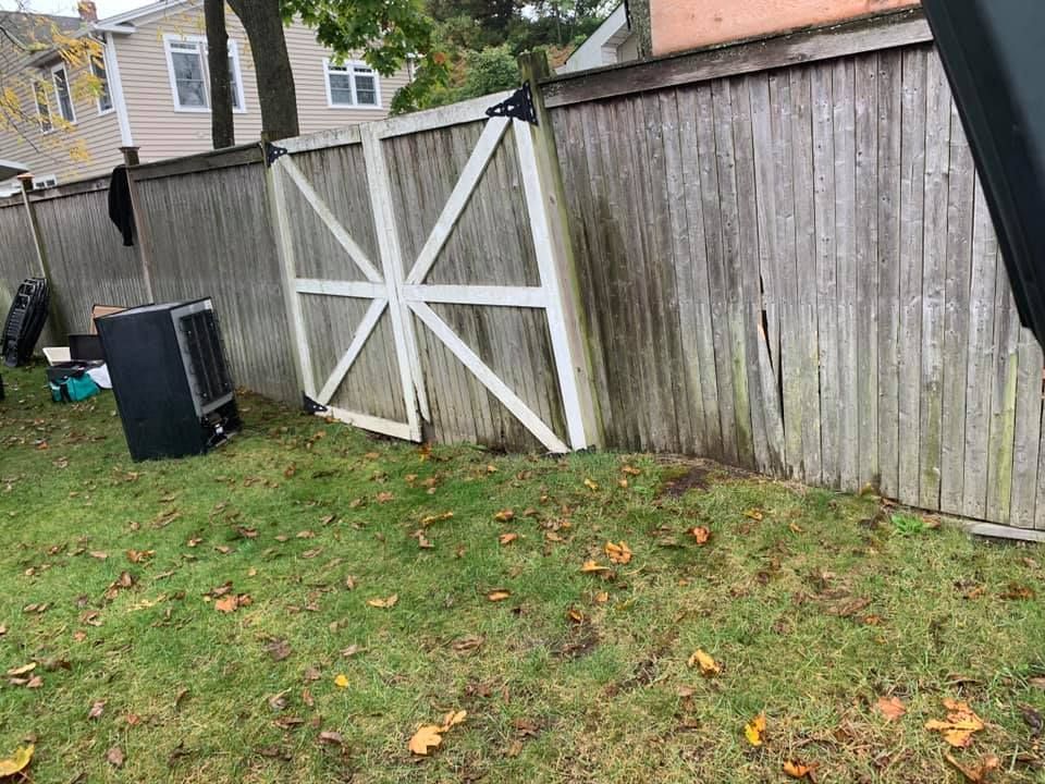 A weathered wooden backyard fence featuring a large, white-trimmed gate, with a black portable cooler on the grass nearby.