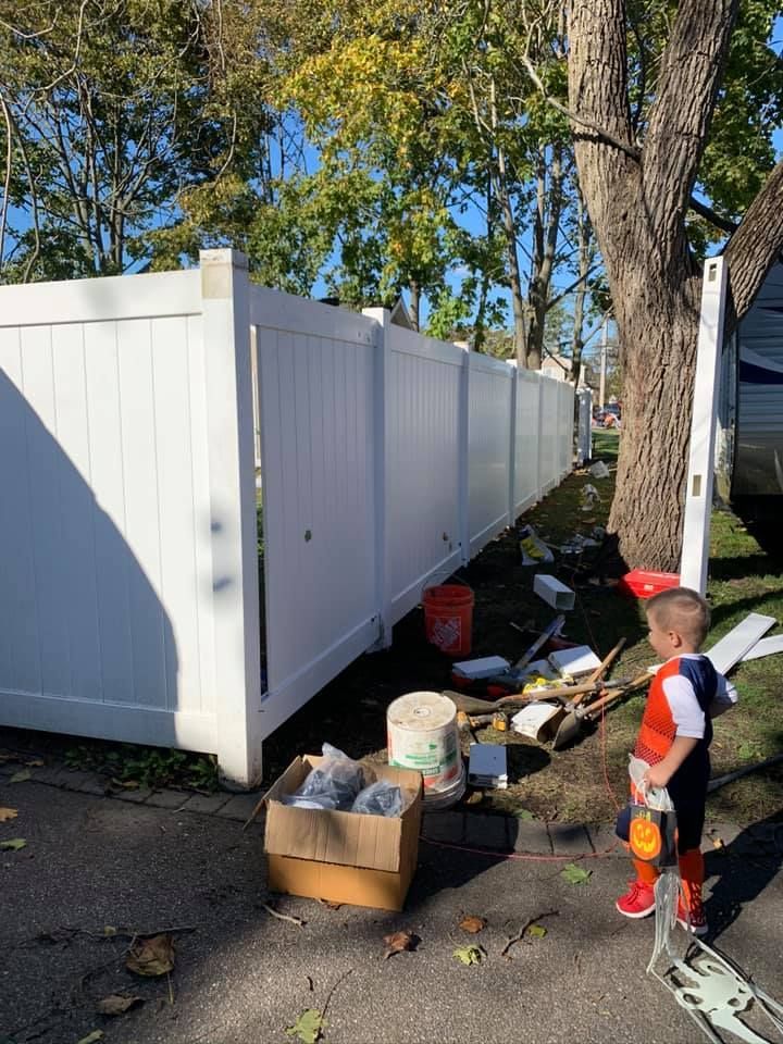 A young child stands near a long, white vinyl privacy fence under construction in a yard with scattered building supplies.