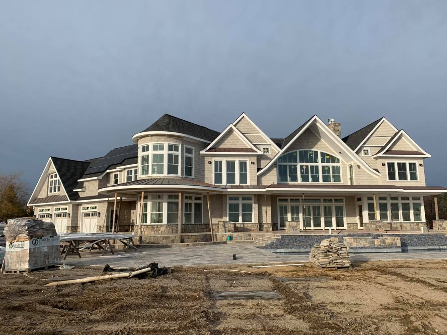 A large, tan, multi-gabled house with stone accents and a partially constructed patio under a cloudy sky.