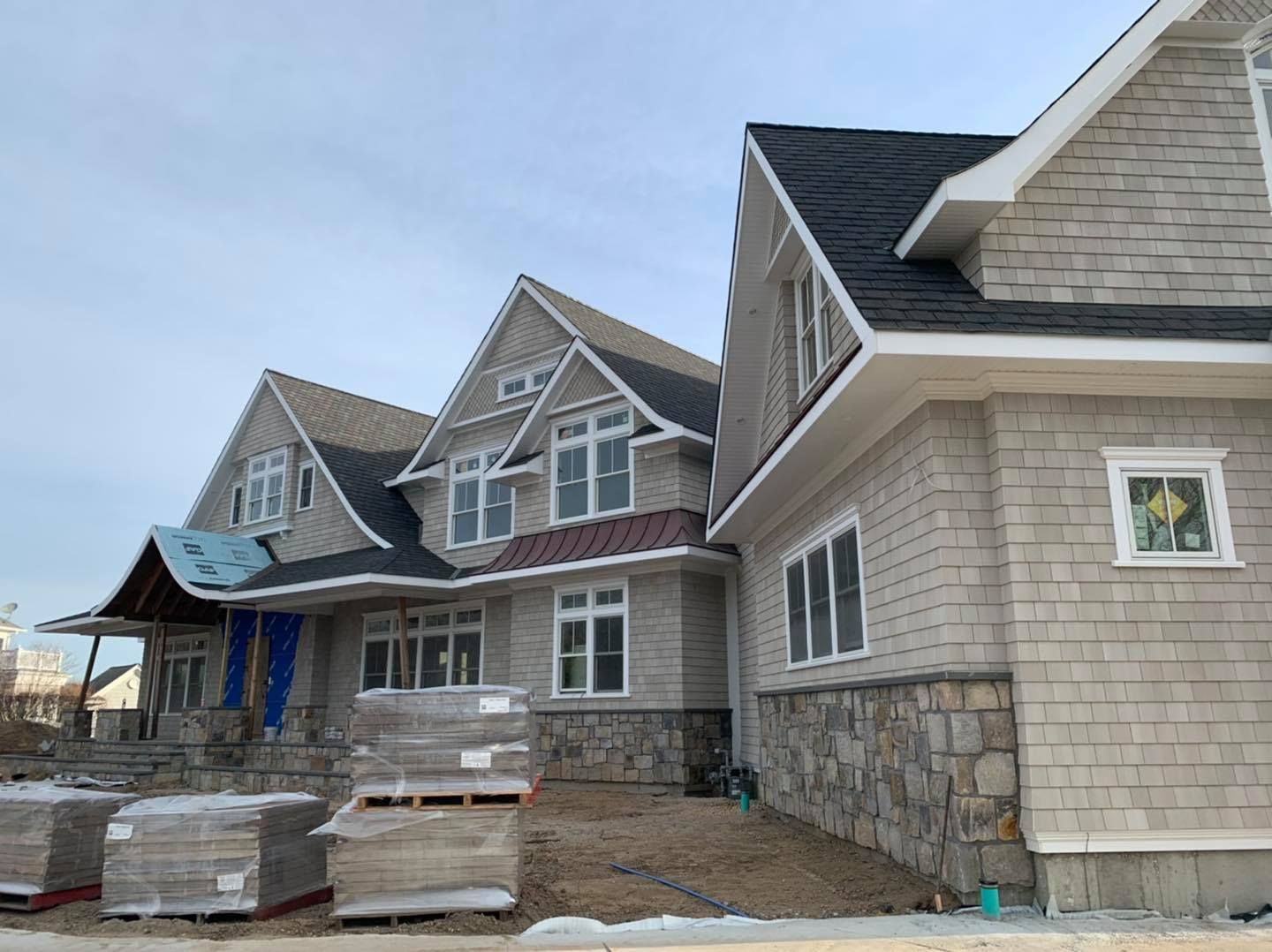 A large, unfinished house with gray shingles and stone siding under construction, with pallets of materials in the yard.