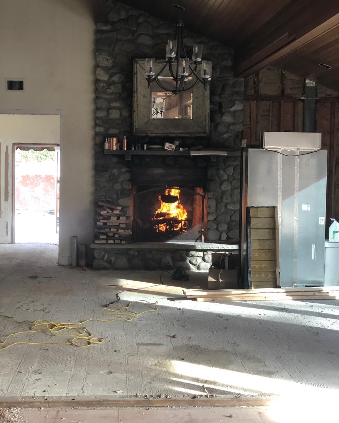 A stone fireplace with a burning fire is in the center of a rustic, unfinished room with an open doorway and wood ceiling.