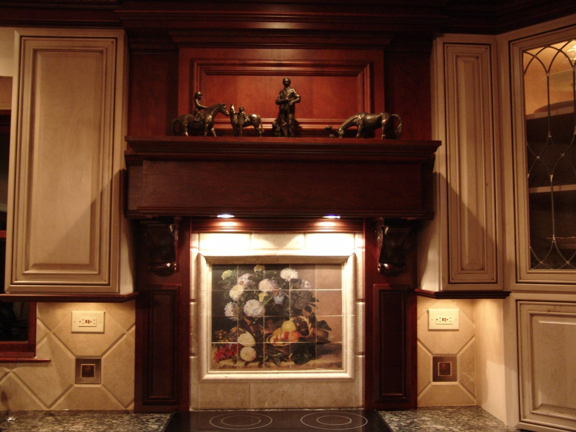 Kitchen stove hood with dark wood, decorative figures on top, and a tiled floral mural backsplash between cabinets.