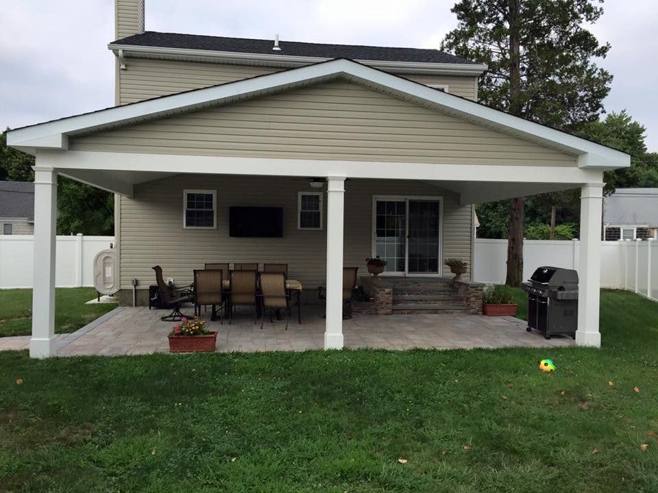 A beige house with a large, covered back patio featuring white pillars, stone flooring, outdoor furniture, and a grill.