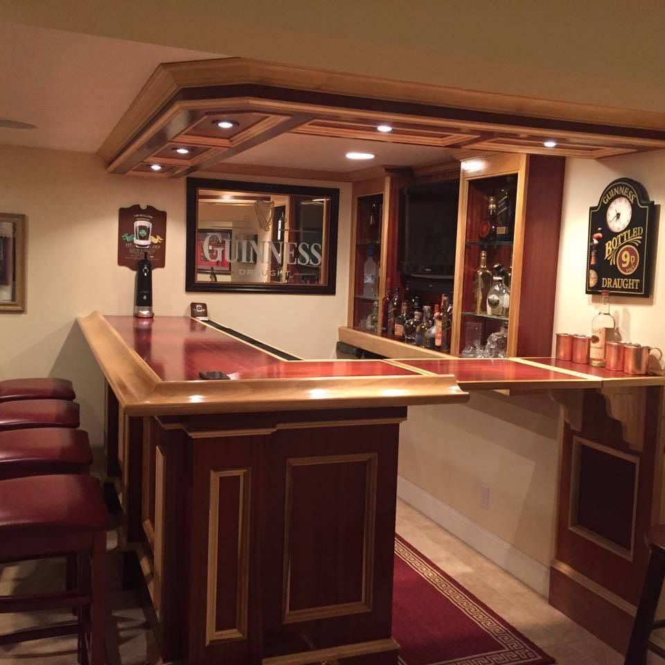 A home basement bar with wooden cabinetry, a dark red countertop, a Guinness mirror, and stools on a red rug.