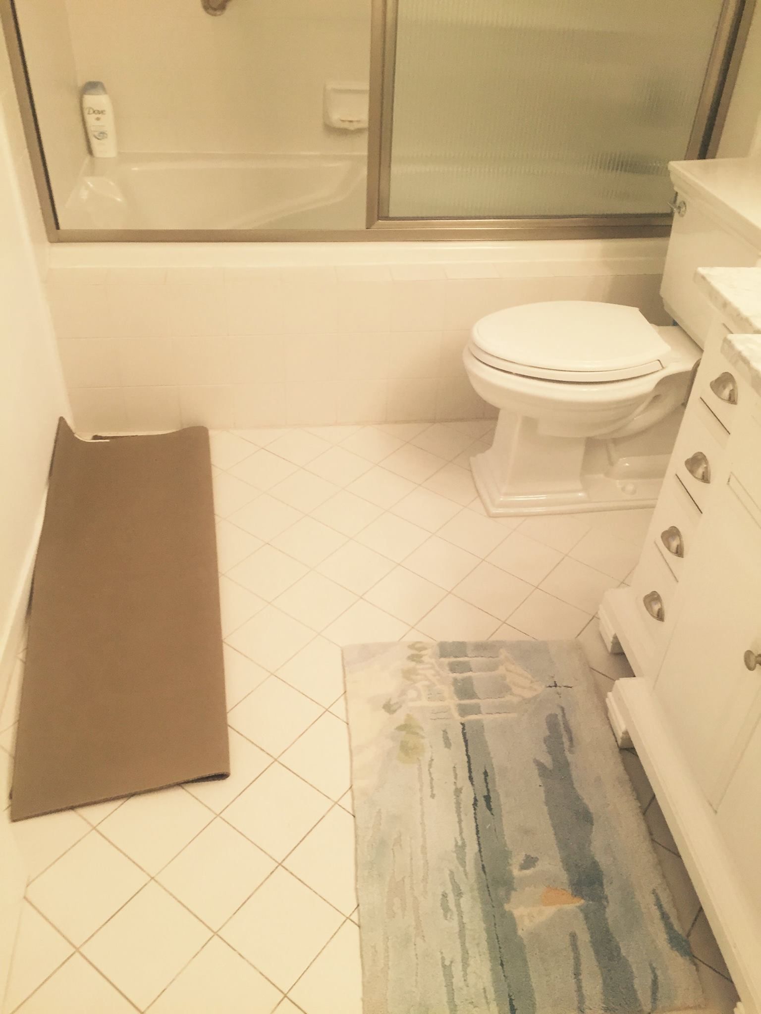 A bathroom with a white toilet, a bathtub, and two rugs, one gray and one patterned, on the white tiled floor.