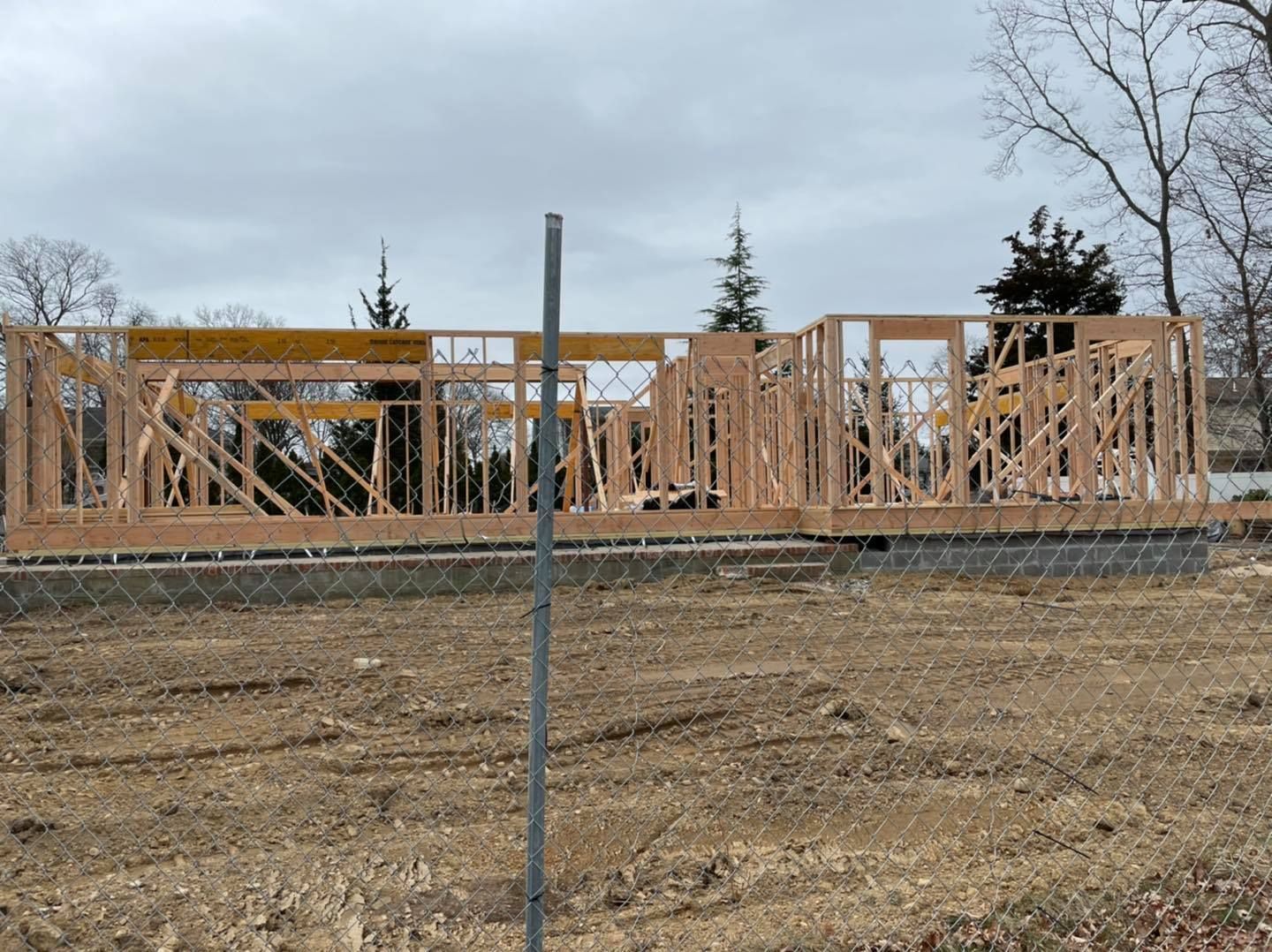 The wooden frame of a new house construction project stands on a concrete foundation in a dirt lot under a cloudy sky.