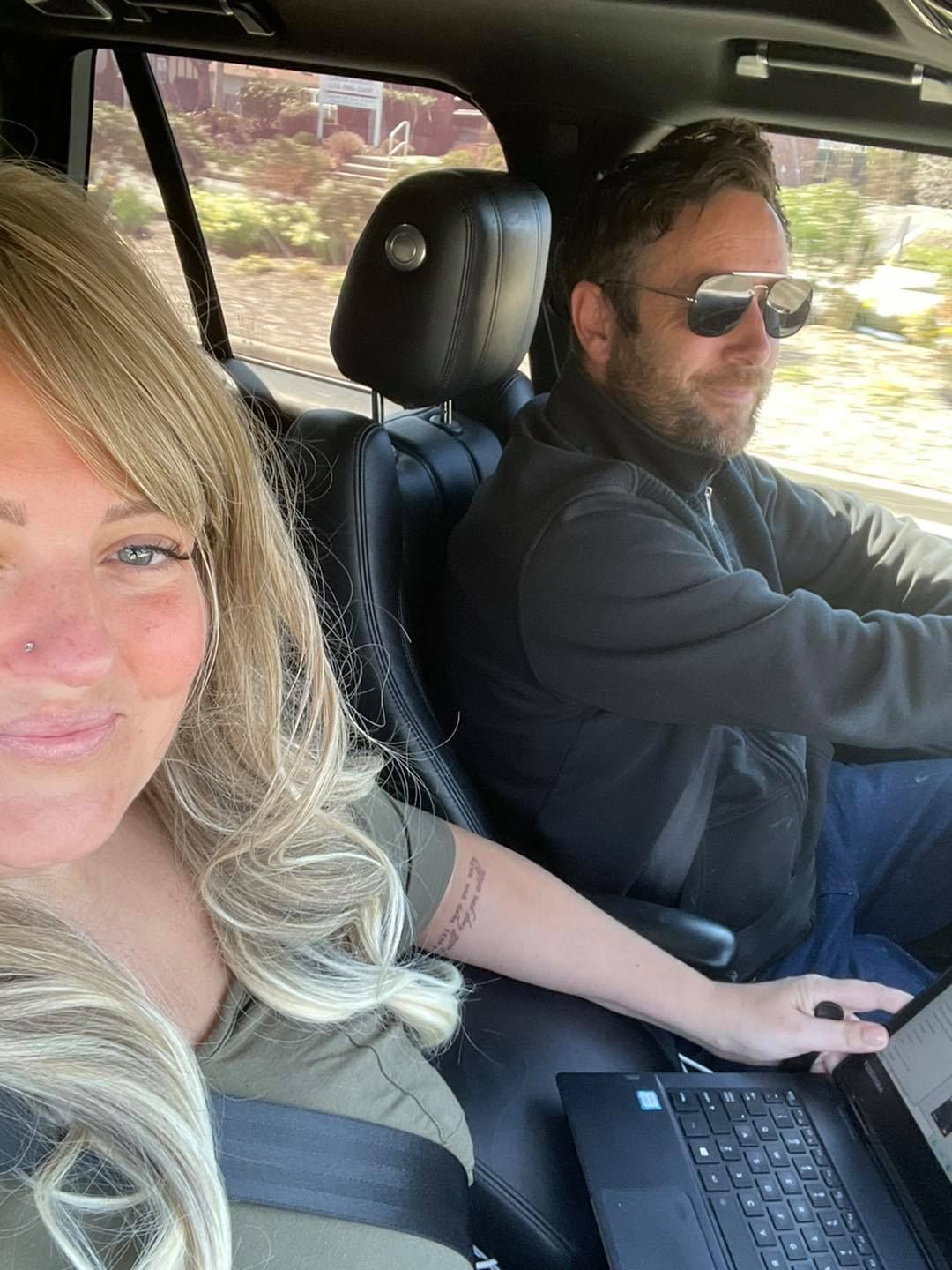A person with blonde hair sits in the passenger seat of a car using a laptop, while another person drives.