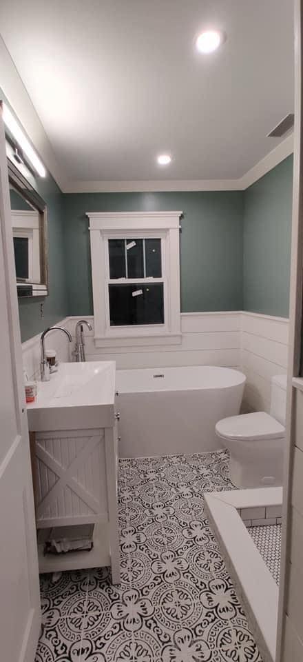 Small bathroom with patterned tile floors, white vanity, freestanding tub, and sage green walls with white wainscoting.