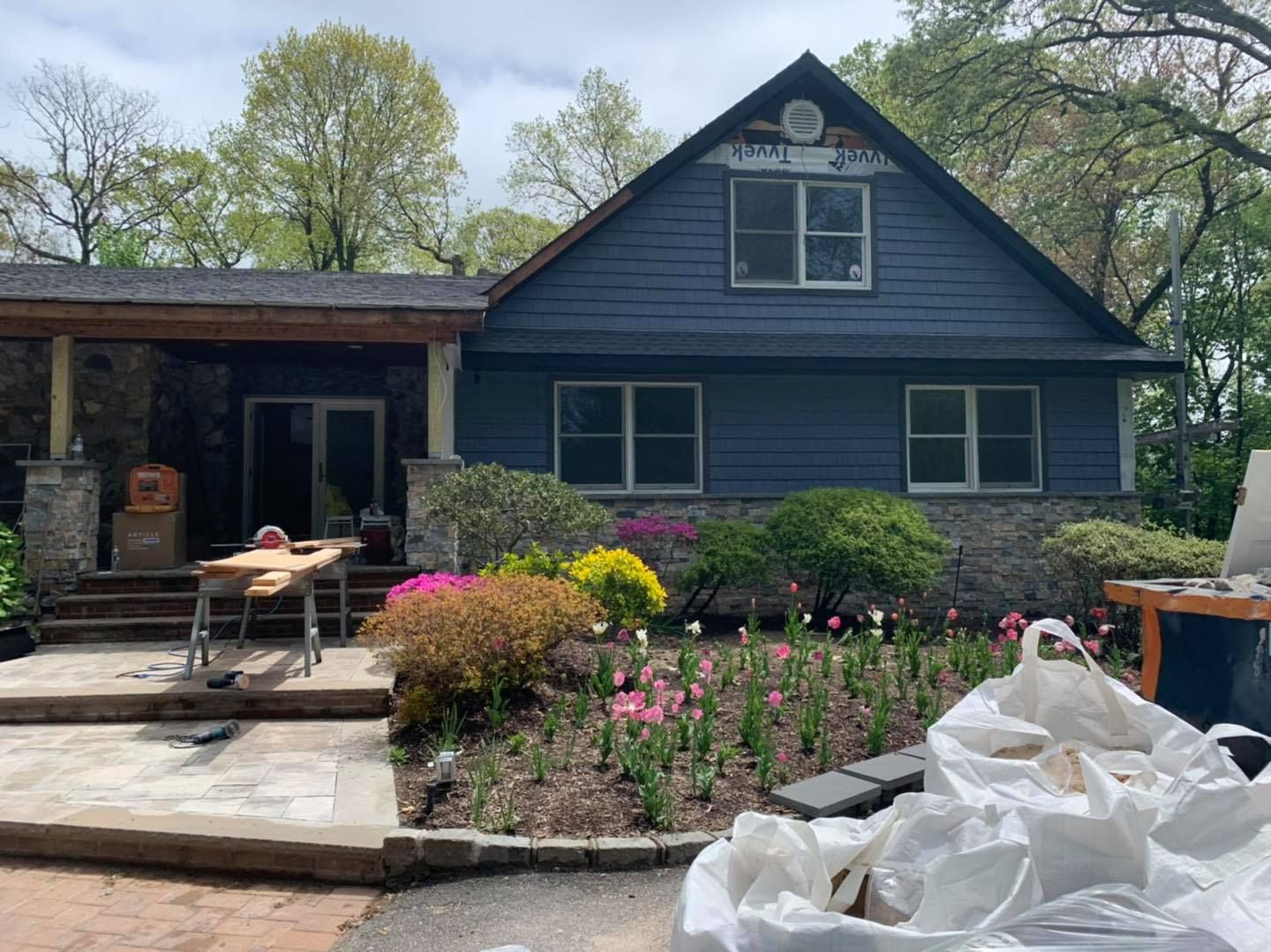 A blue house with a stone base features a landscaped front yard with shrubs and flowers, and construction supplies.