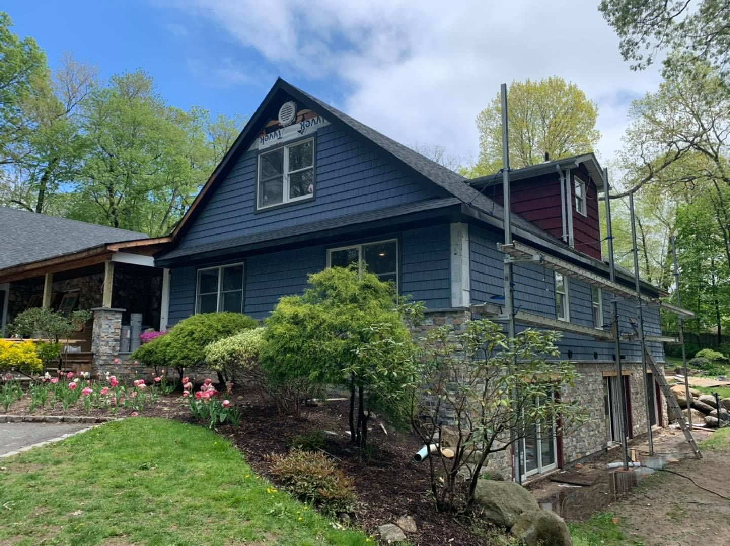 A two-story blue house with stone siding on the lower level, surrounded by trees, garden landscaping, and scaffolding.