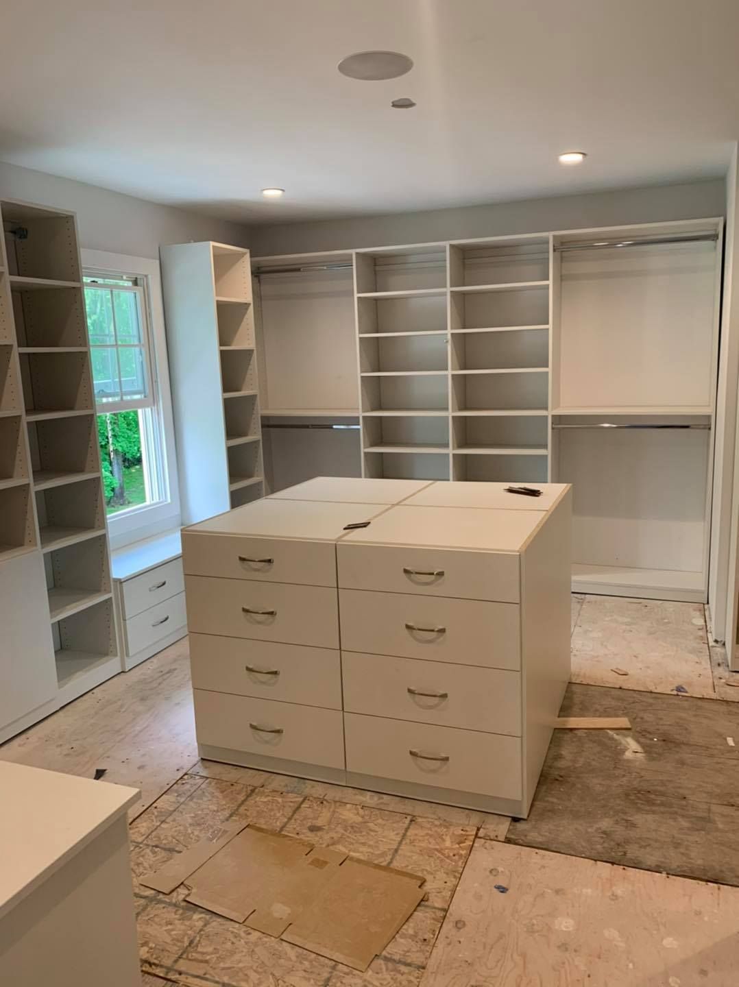 A white closet installation under construction, featuring wall-to-wall shelving, hanging rods, and a central dresser unit.