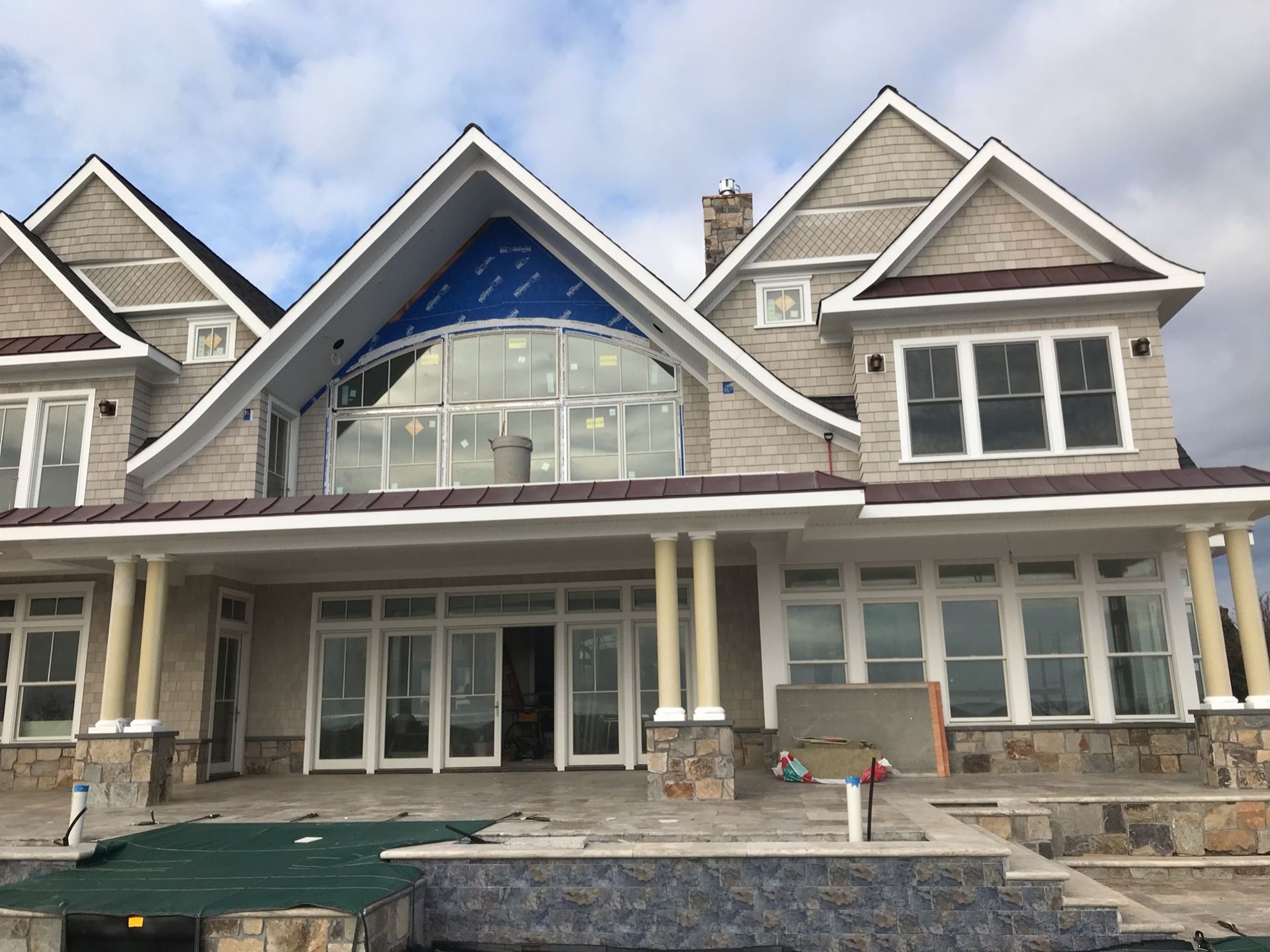 A large, multi-story stone house with a covered patio, arched windows, and a blue protective film on a gable window.