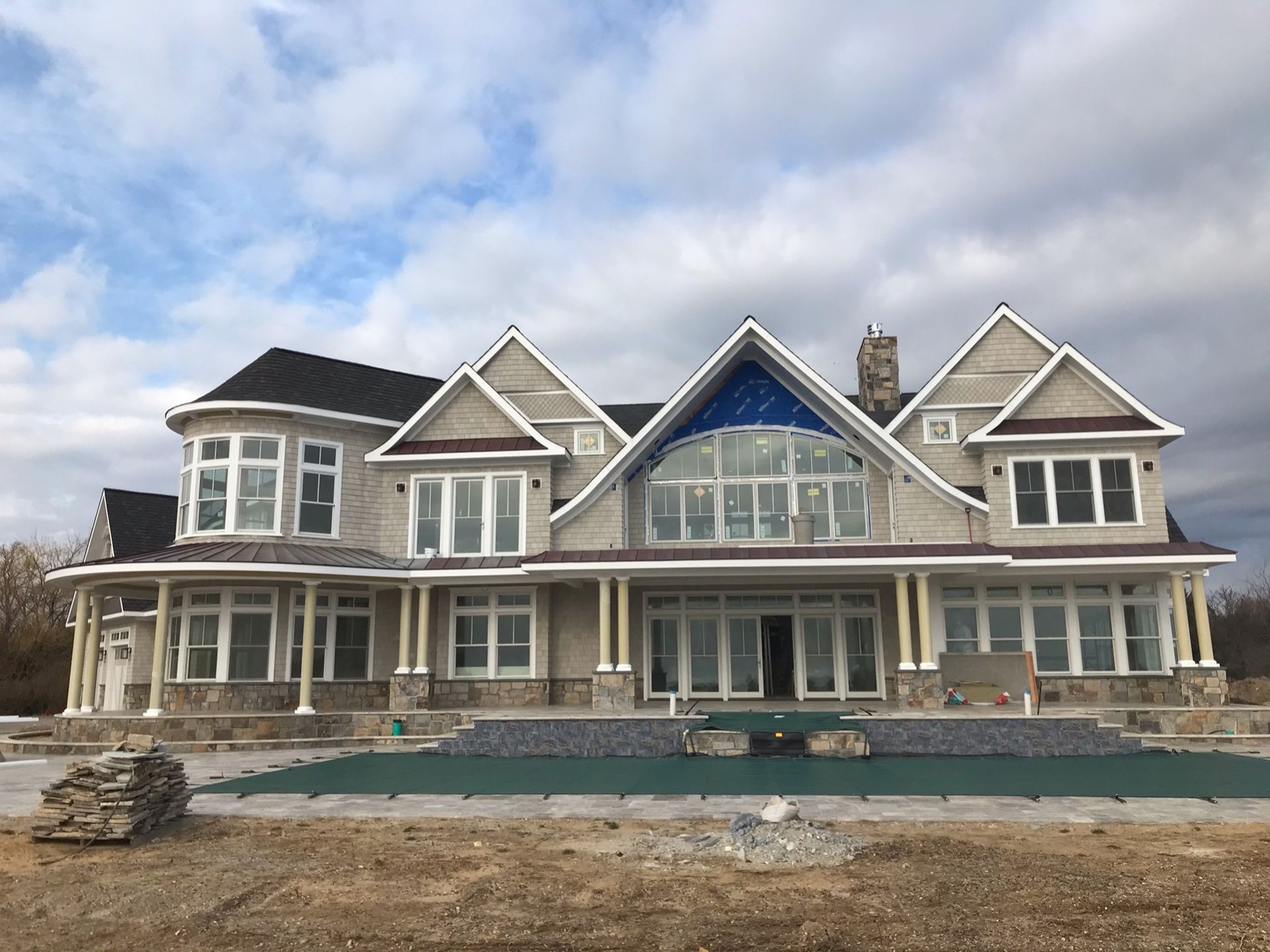 A large, multi-story stone house under construction with a covered swimming pool in the foreground.