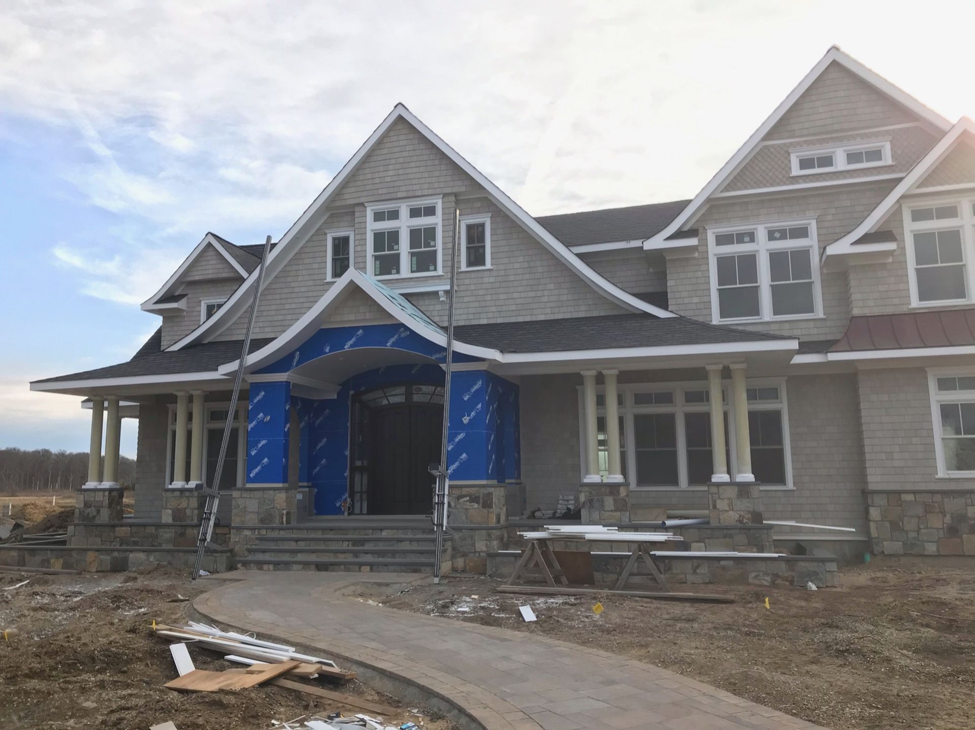 A large, multi-gabled house under construction with tan shingles, stone accents, and a curved stone walkway.