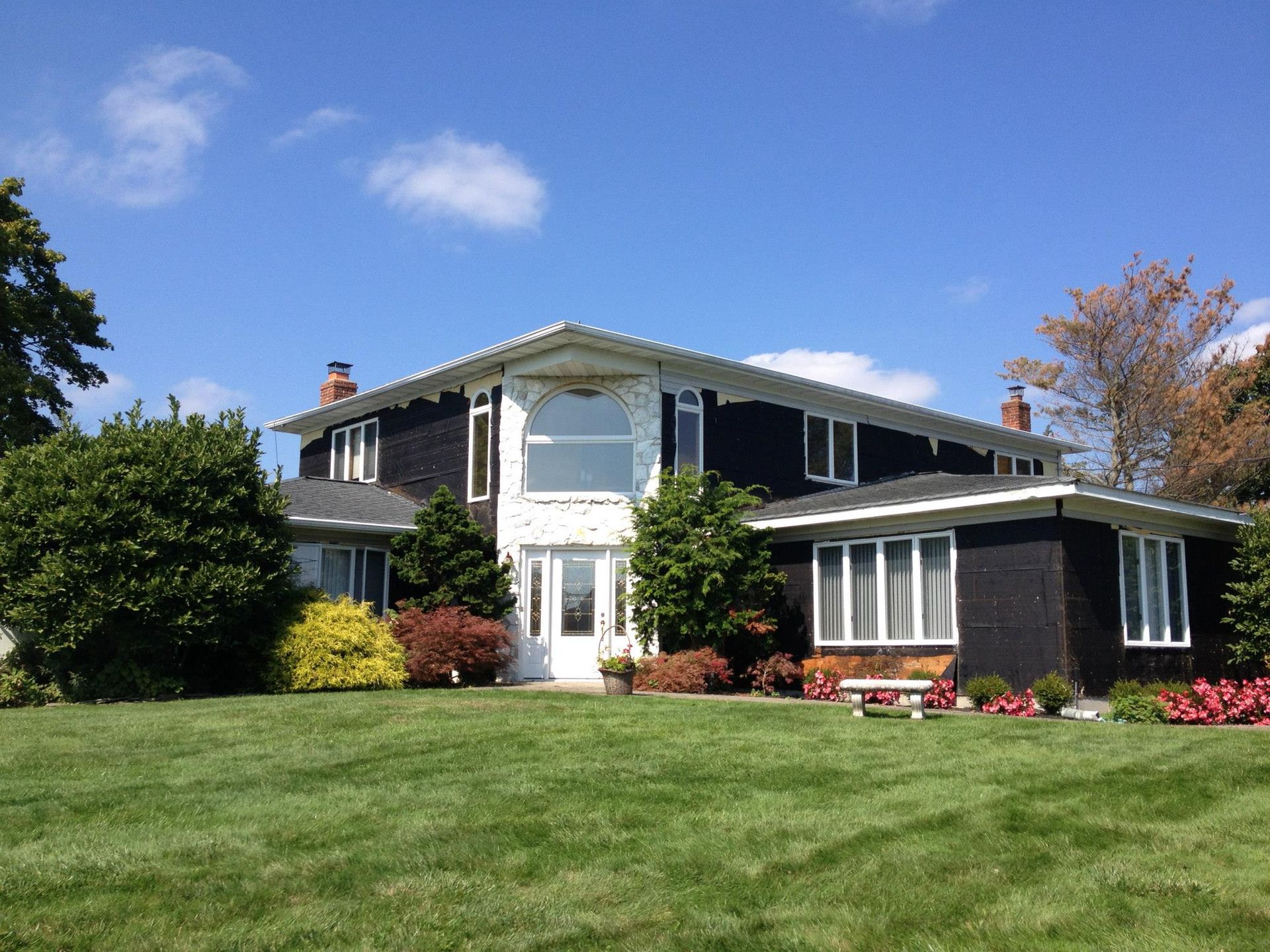A large, two-story house with dark siding and white trim, set behind a lush green lawn under a bright blue sky.