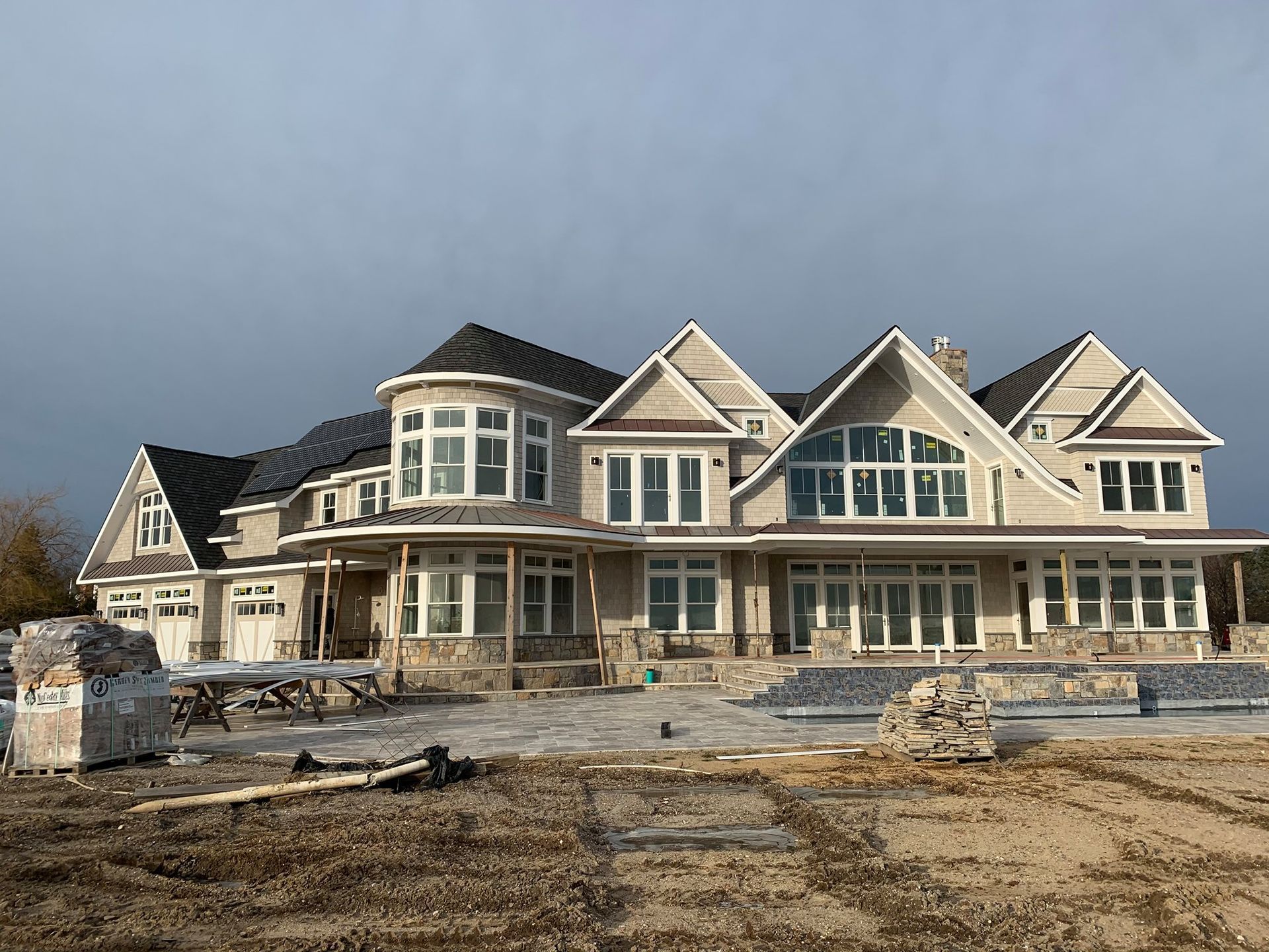 Large, multi-story house under construction with tan siding, stone base, and dark gabled roofs on a dirt lot.