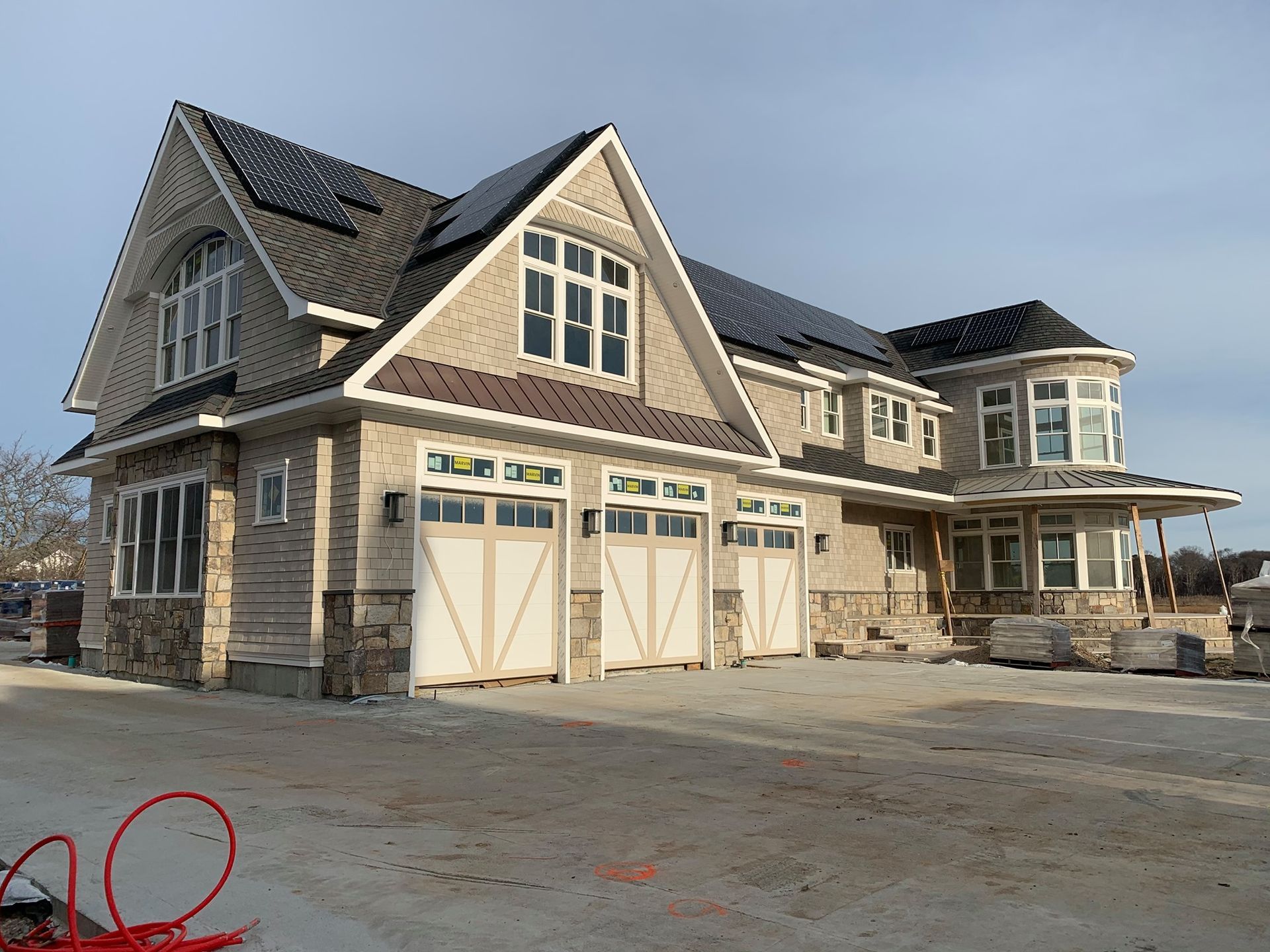 A large, multi-story suburban home under construction with a three-car garage, stone facade, and dark shingled roof.
