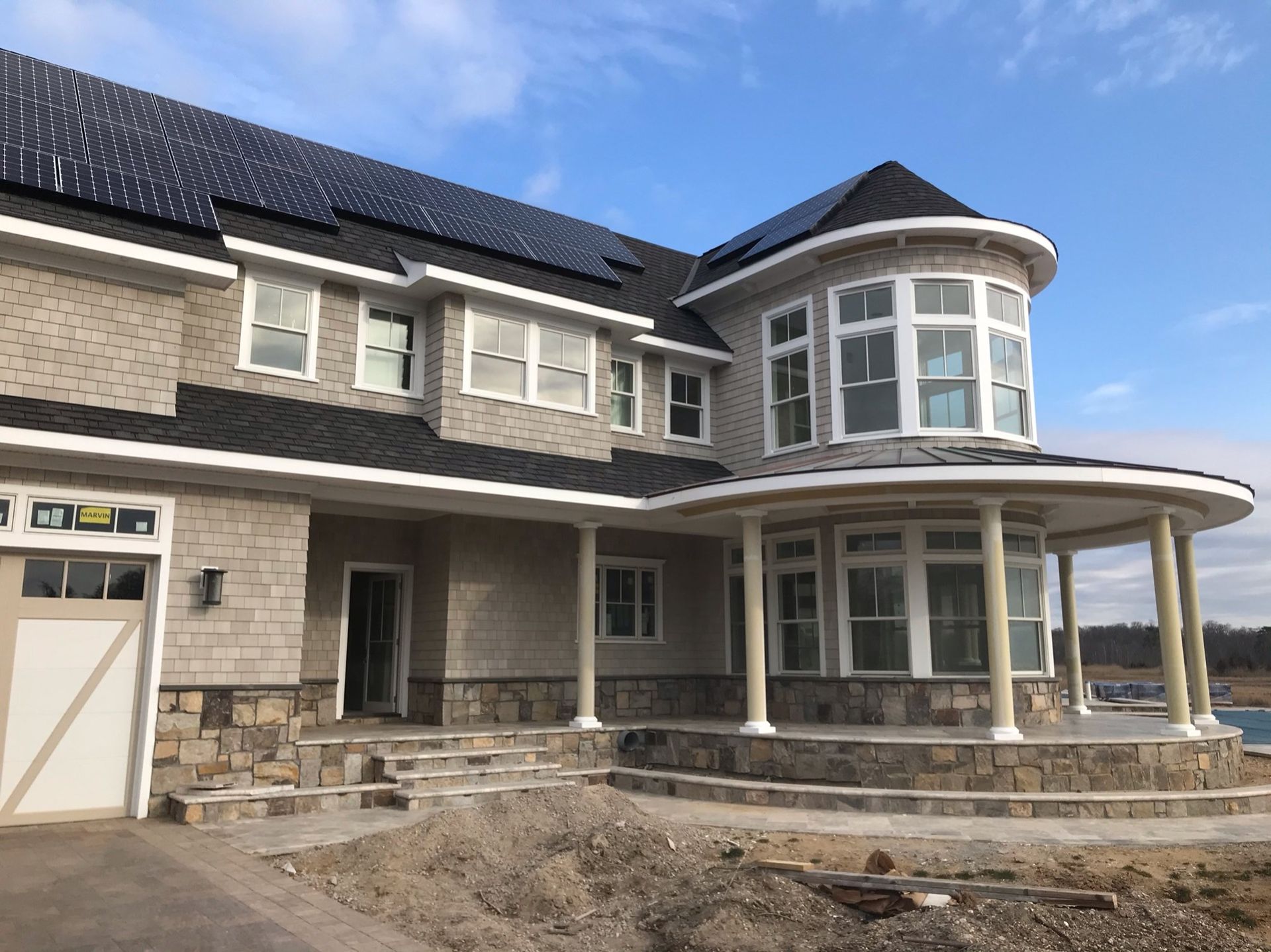 A partially constructed two-story house with a round tower, gray siding, stone foundation, and black solar roof panels.