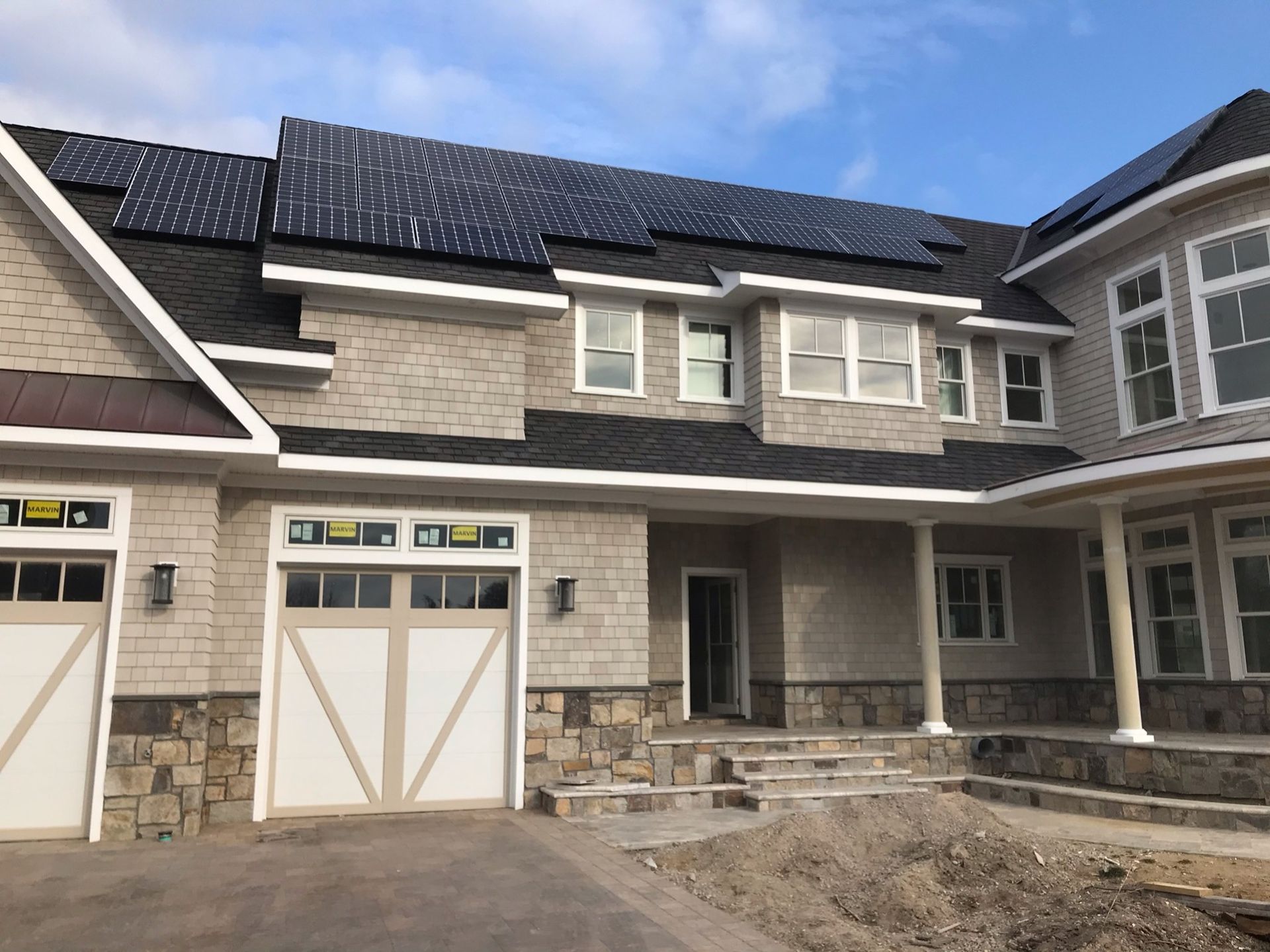 A large, multi-story house under construction with stone siding, a shingled roof with solar panels, and garage doors.