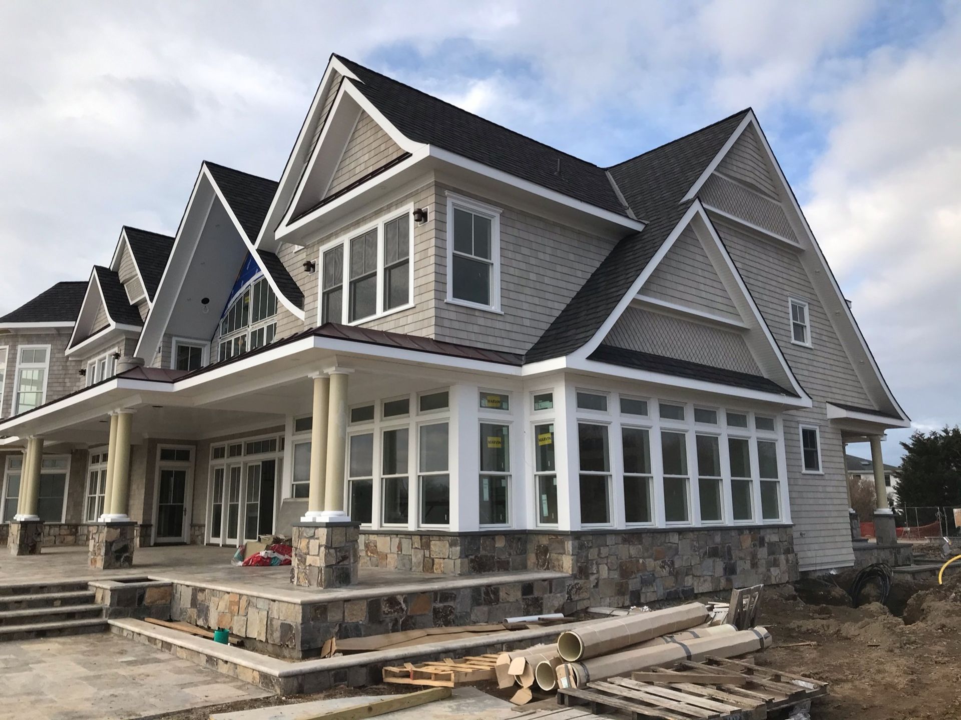A partially finished two-story house with gray shingle siding, a stone foundation, a wrap-around porch, and black roof.