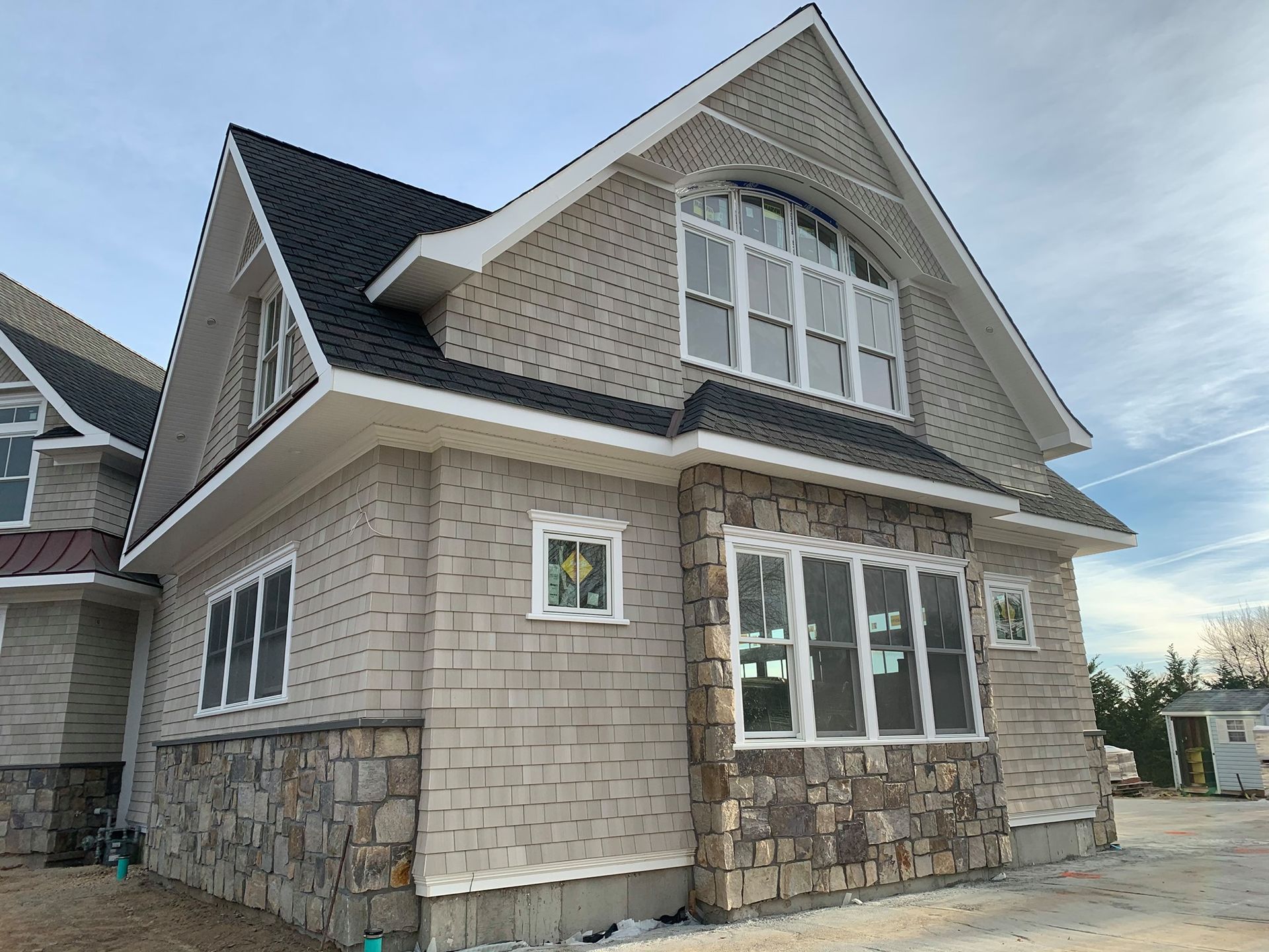 A tan house under construction with stone-veneer siding on the lower level, a dark roof, and a large arched window.