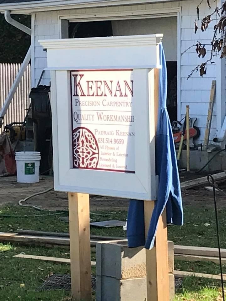 A wooden sign for Keenan Precision Carpentry stands in front of a house under construction with a blue shirt hanging on it.
