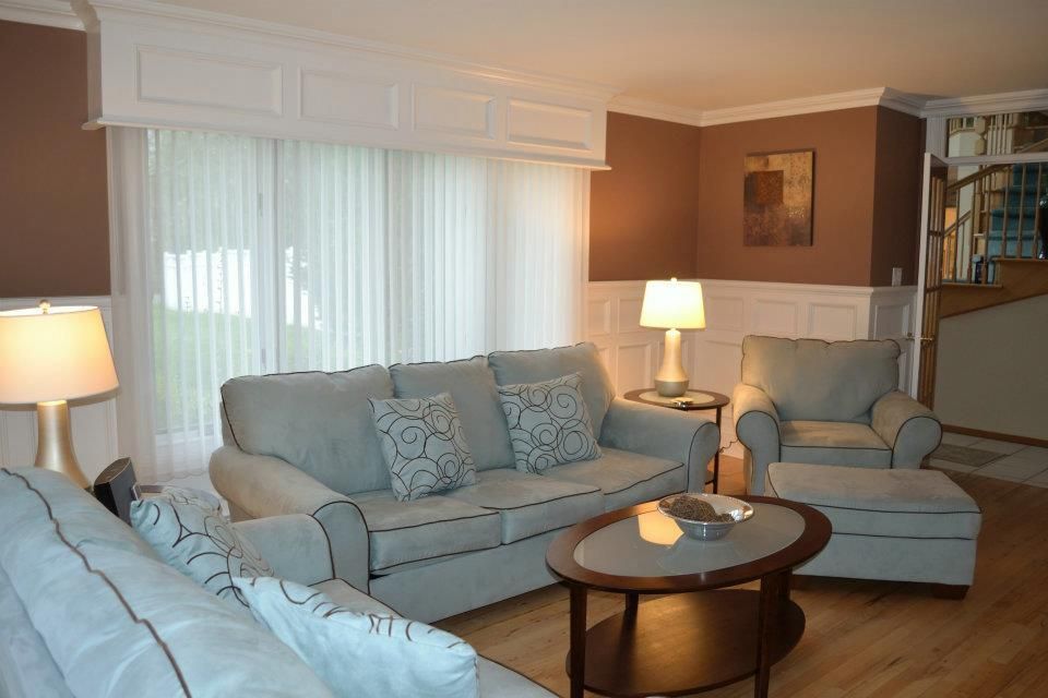 A living room with light blue sofas, a round coffee table, and an armchair against brown walls and white wainscoting.