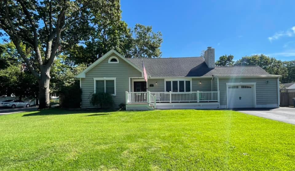A sage green, single-story house with a front porch, an attached garage, and a large front lawn under a clear blue sky.