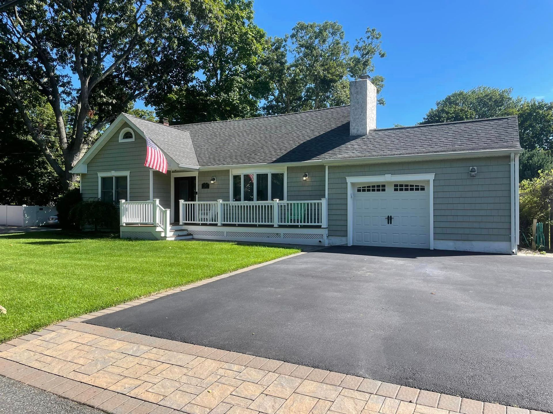 A light gray, single-story house with a white front porch, attached garage, and a paved driveway under a clear blue sky.