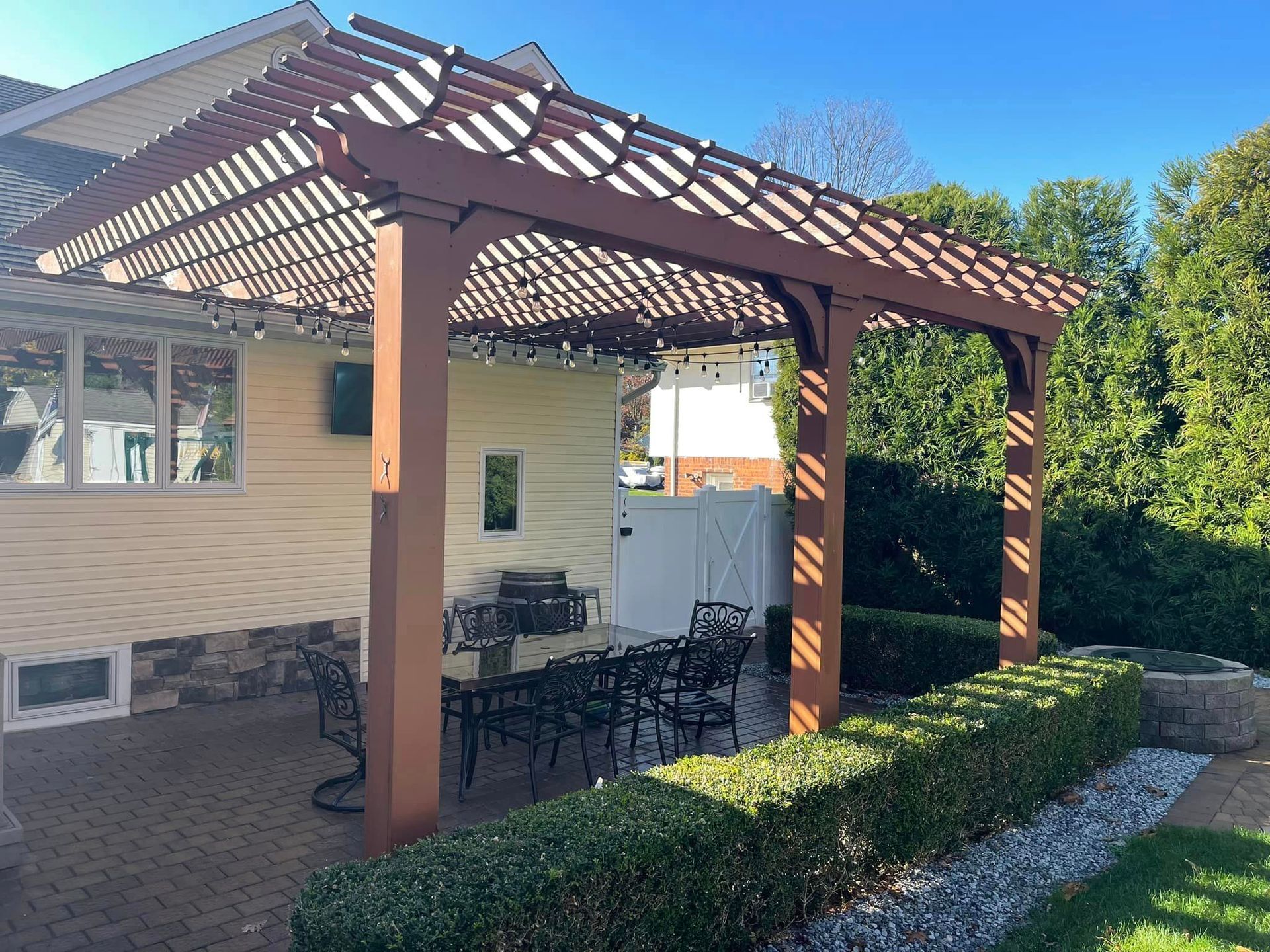 A wooden pergola covers a patio dining set next to a tan house, with a manicured green hedge along the side.