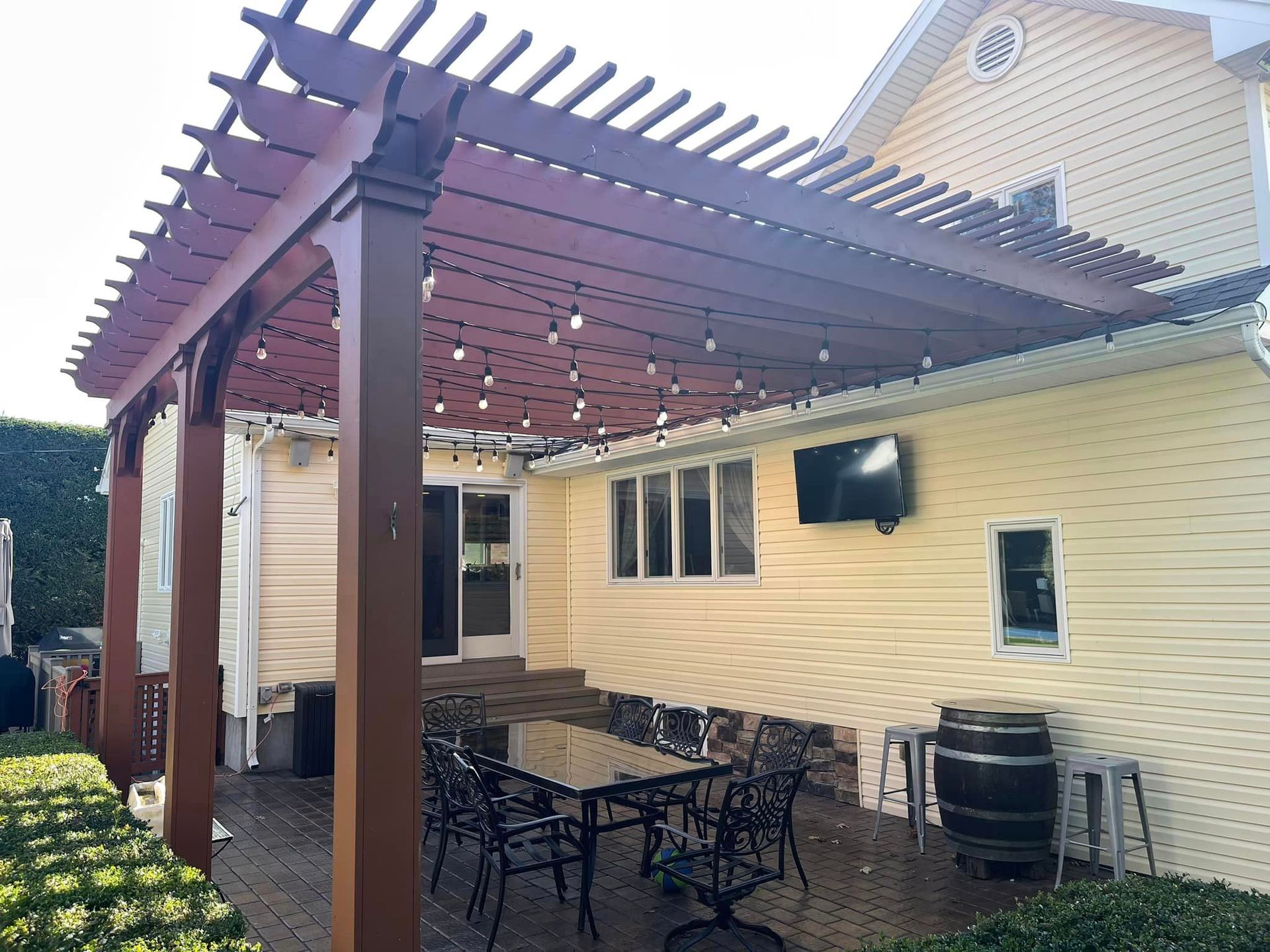 A wooden pergola over a patio with a dining set, wall-mounted television, and string lights against a yellow house.