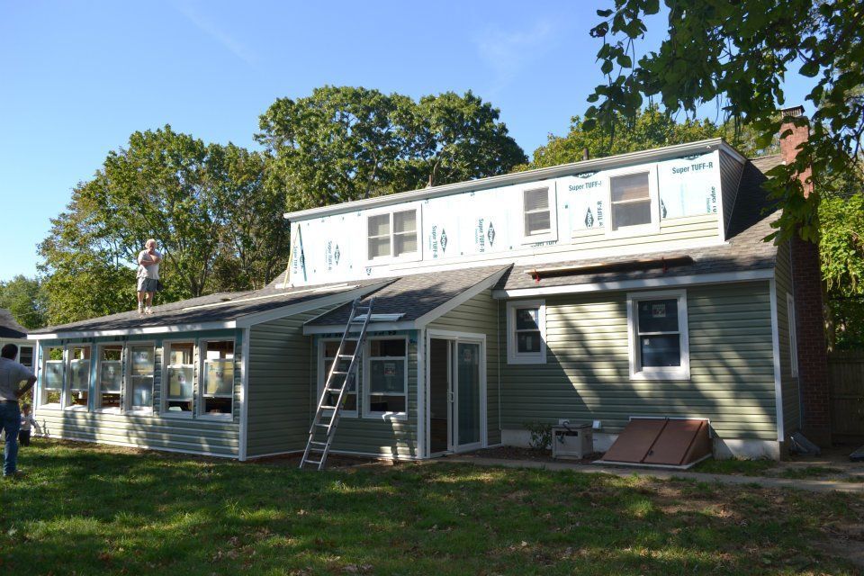 A house under construction with exposed sheathing on the upper level, green siding, and a person on the roof.