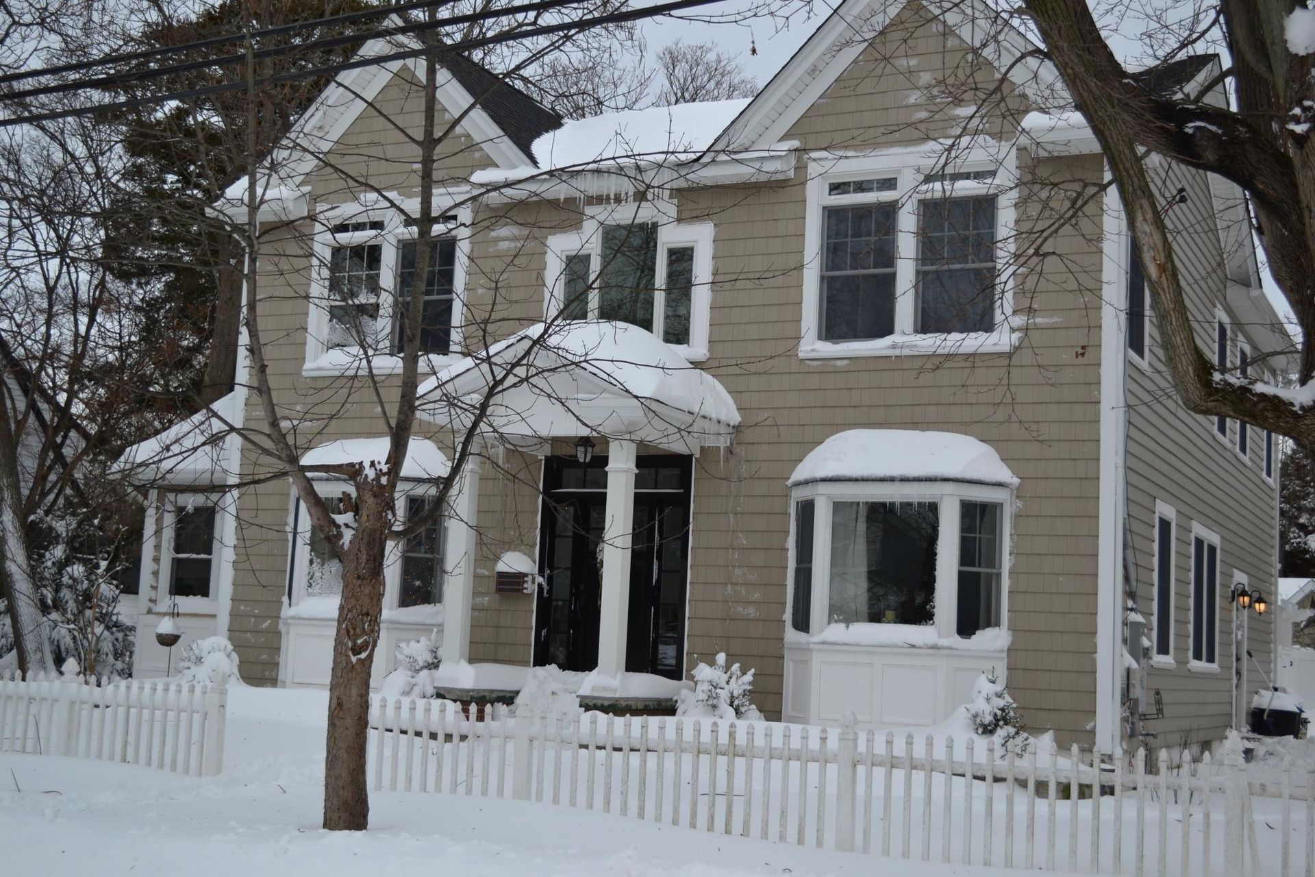 A two-story tan house with a snow-covered yard, a white picket fence, and a front porch, all blanketed in winter snow.