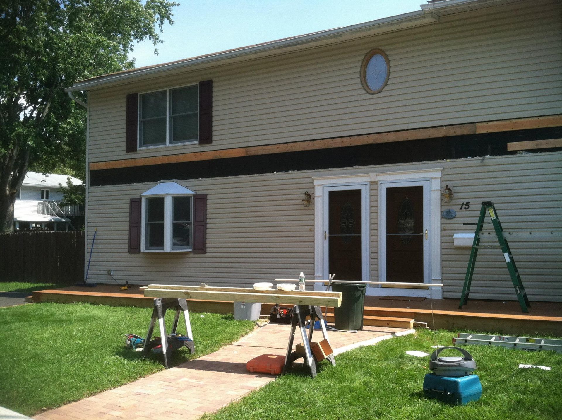 A house exterior under construction with a deck ledger board being installed, sawhorses, and tools on the lawn.