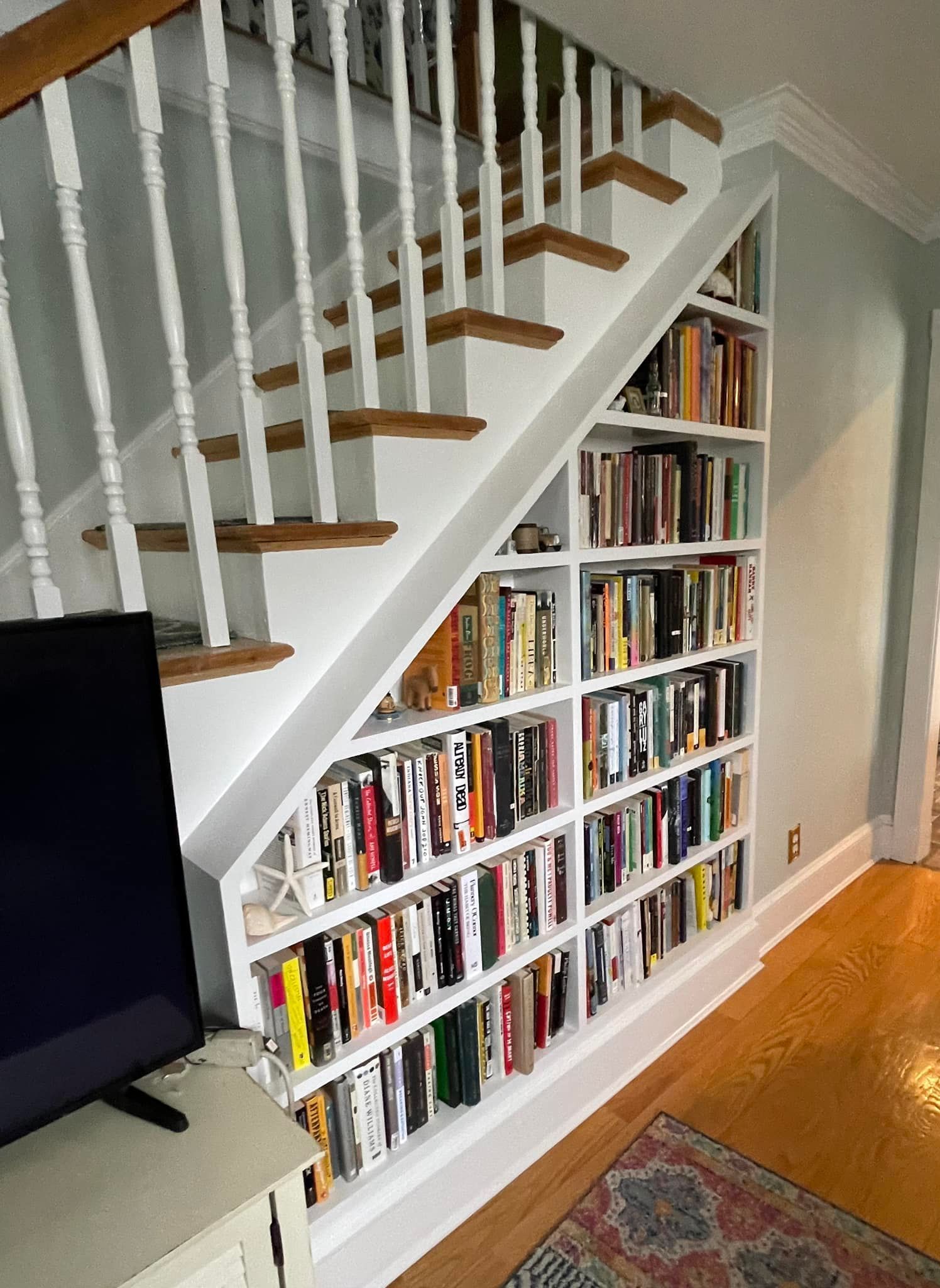 Built-in white bookshelves are tucked under a wooden staircase with white railings, filled with many colorful books.