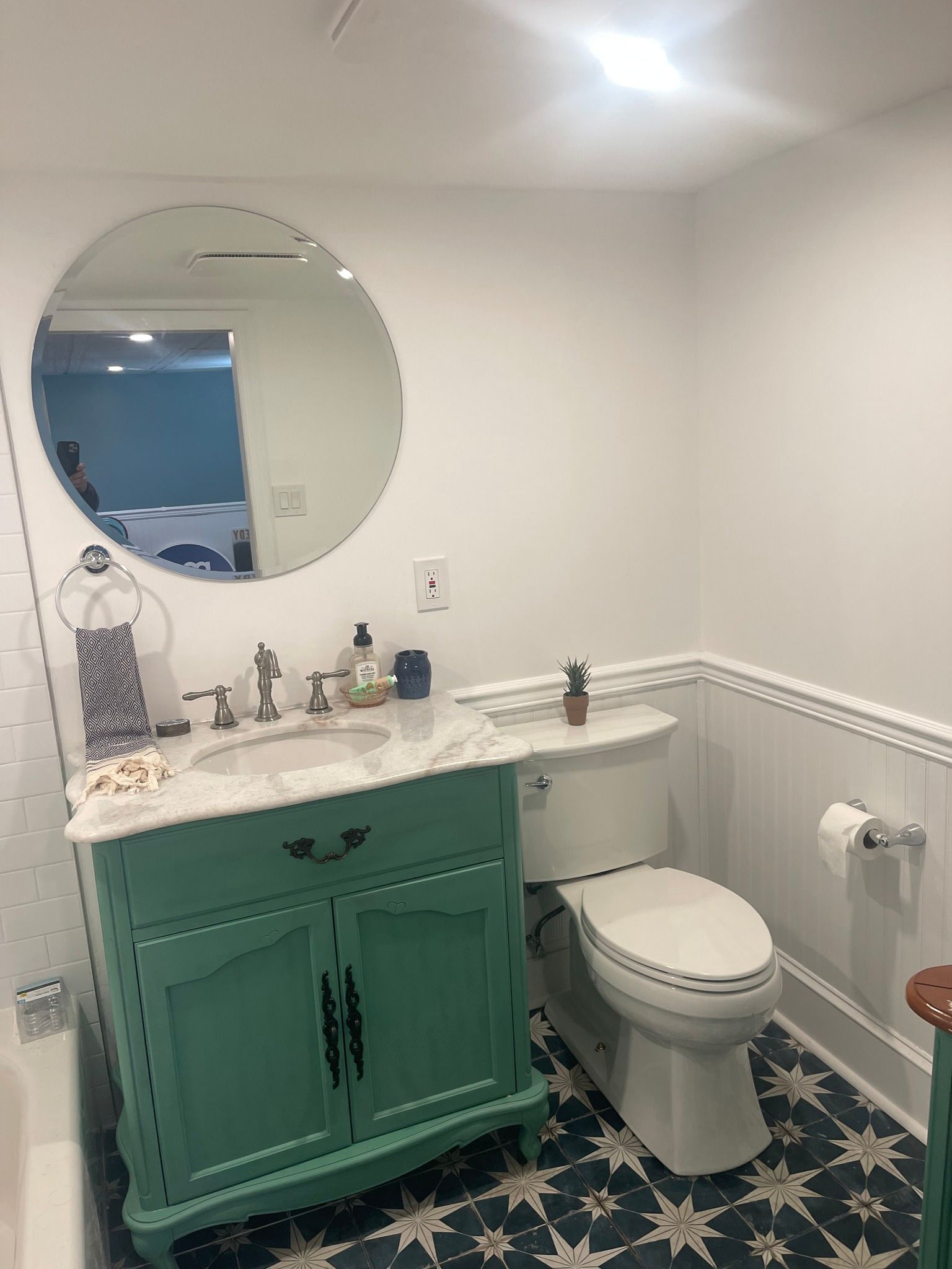 A bathroom featuring a green vanity with a marble top, a round mirror, white walls with wainscoting, and a patterned floor.
