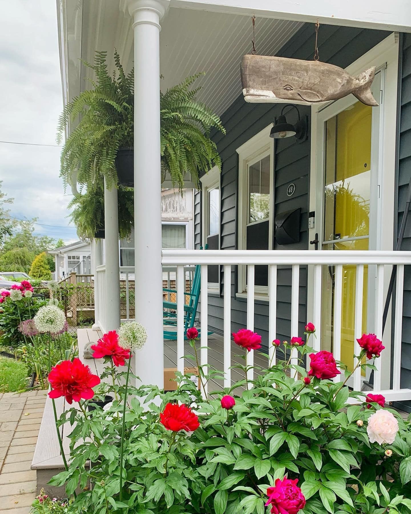A blue house with a porch, hanging ferns, a wooden whale decoration, a yellow front door, and red peonies in the garden.