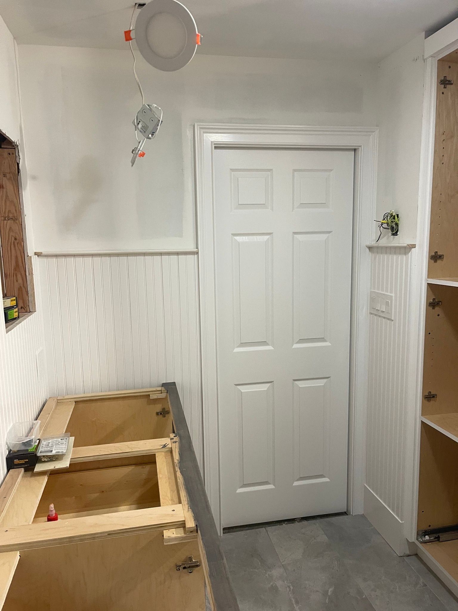 A room under renovation featuring unfinished wooden cabinets, white wainscoting, a white door, and an exposed ceiling light.