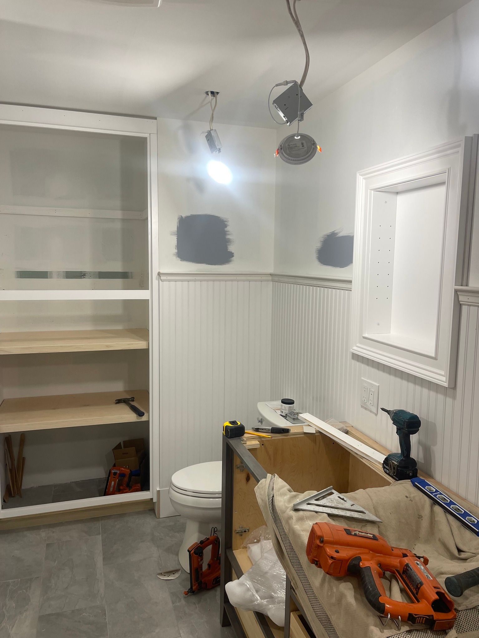 A bathroom under construction with wall panels, a toilet, an open shelving unit, and tools on a vanity cabinet.