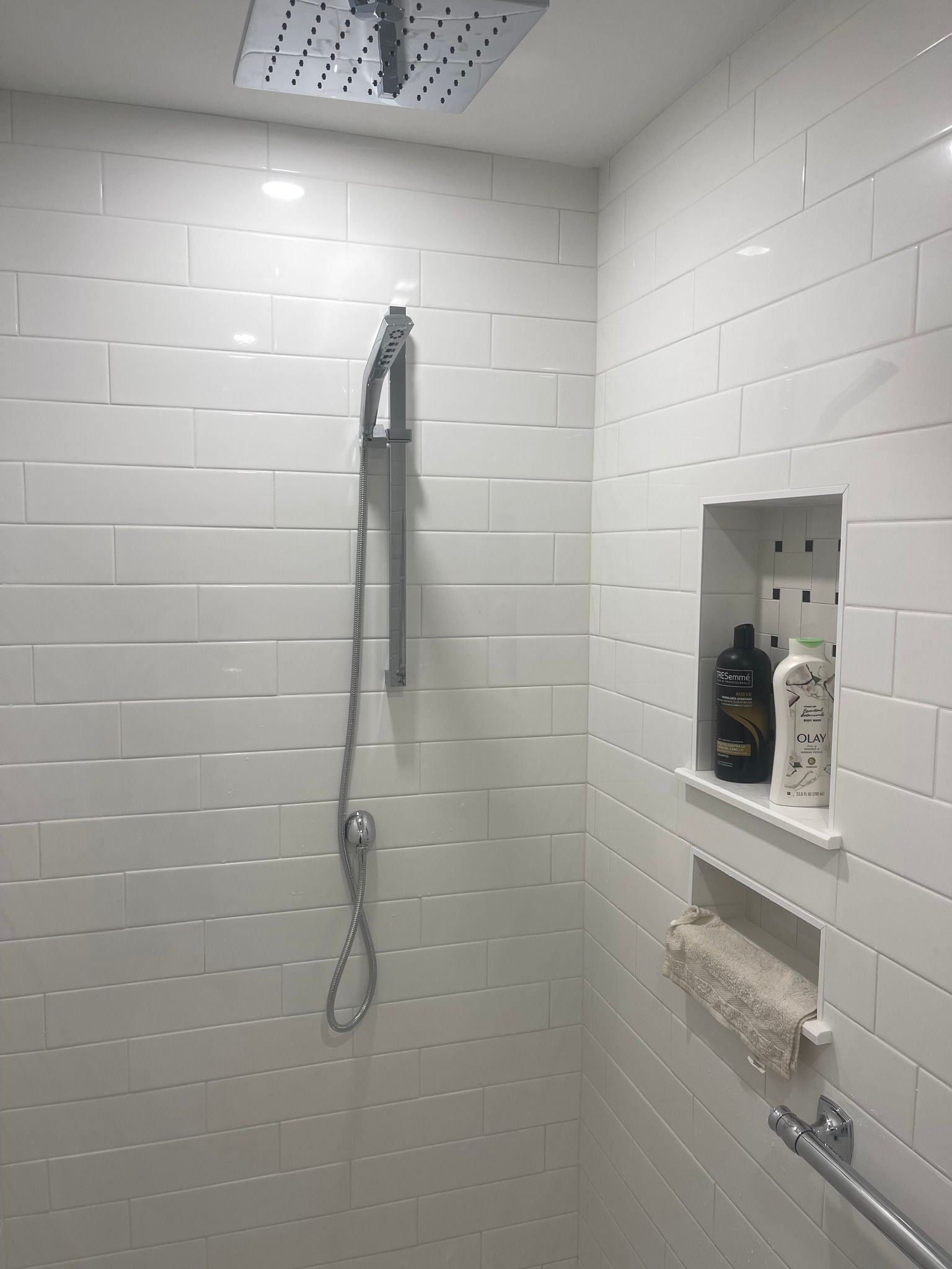 A modern white-tiled shower featuring a rainfall showerhead, a handheld spray, and two recessed shelves holding soap.