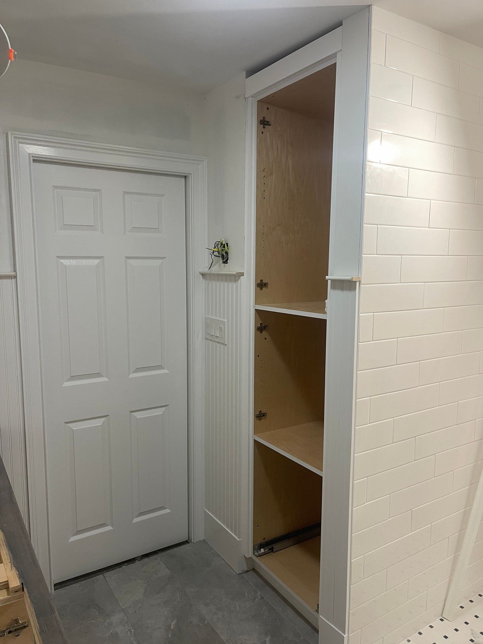 A white door next to an open, unfinished wooden pantry cupboard set against a white brick-style wall.