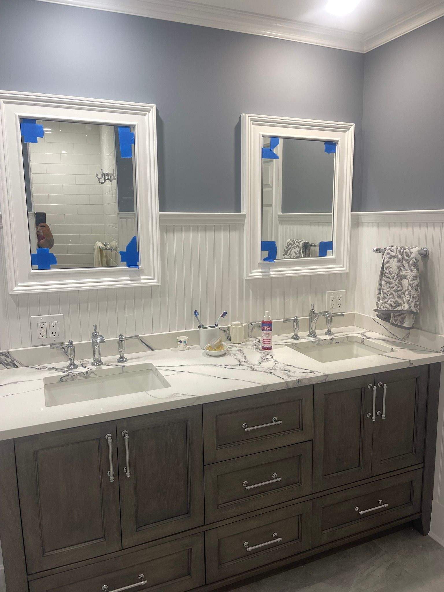 A double bathroom vanity with grey cabinets, a white countertop, two sinks, and white-framed mirrors on a grey wall.